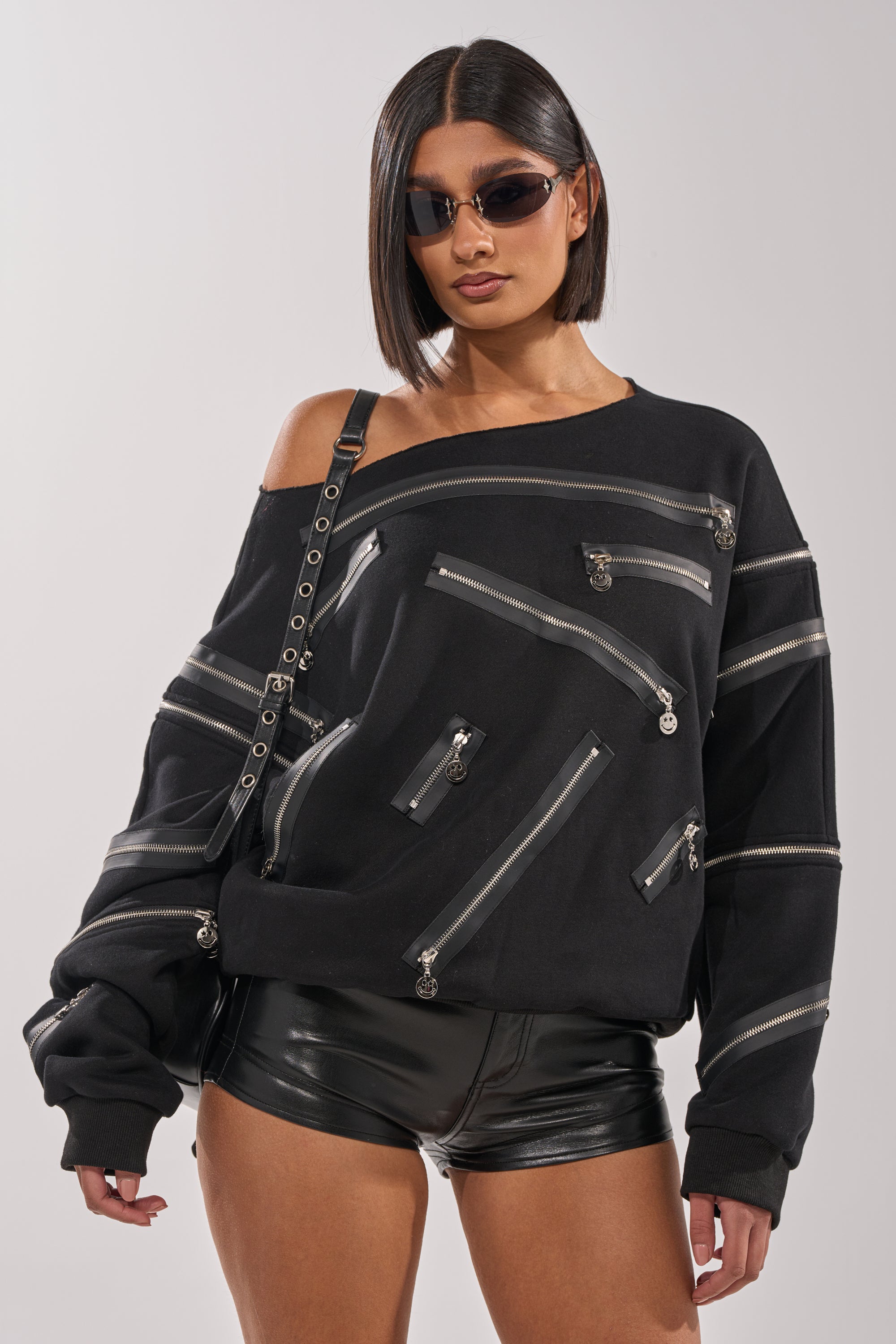 A woman with a sleek bob and sunglasses models the UN-ZIP ME OFF THE SHOULDER SWEATSHIRT, featuring silver zippers, styled streetwear with black leather shorts against a plain light background. 2