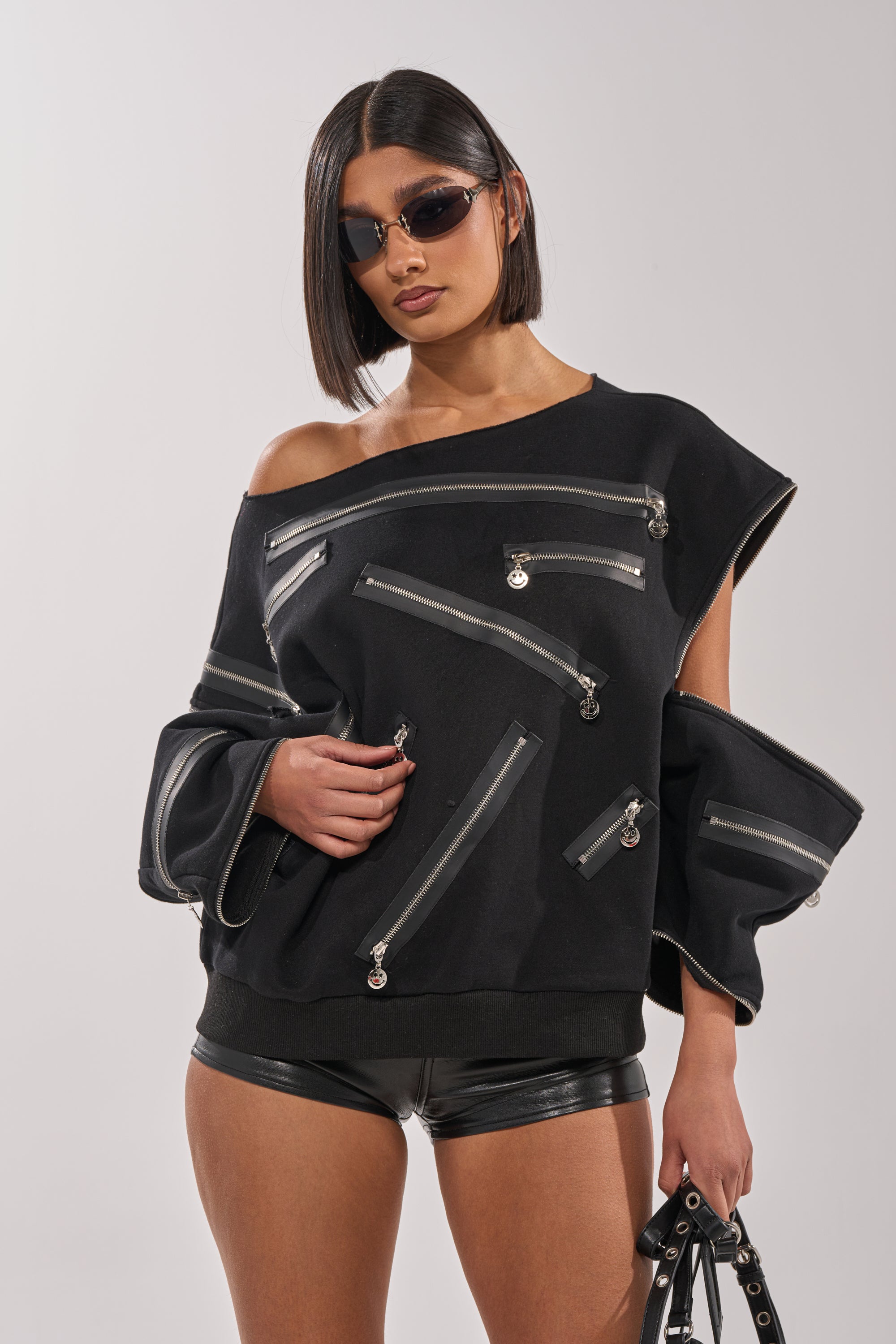 A woman with a sleek bob wears sunglasses and the UN-ZIP ME OFF THE SHOULDER SWEATSHIRT, featuring diagonal zippers, paired with black leather shorts. She carries a black bag with metal rings and stands against a plain background. 4