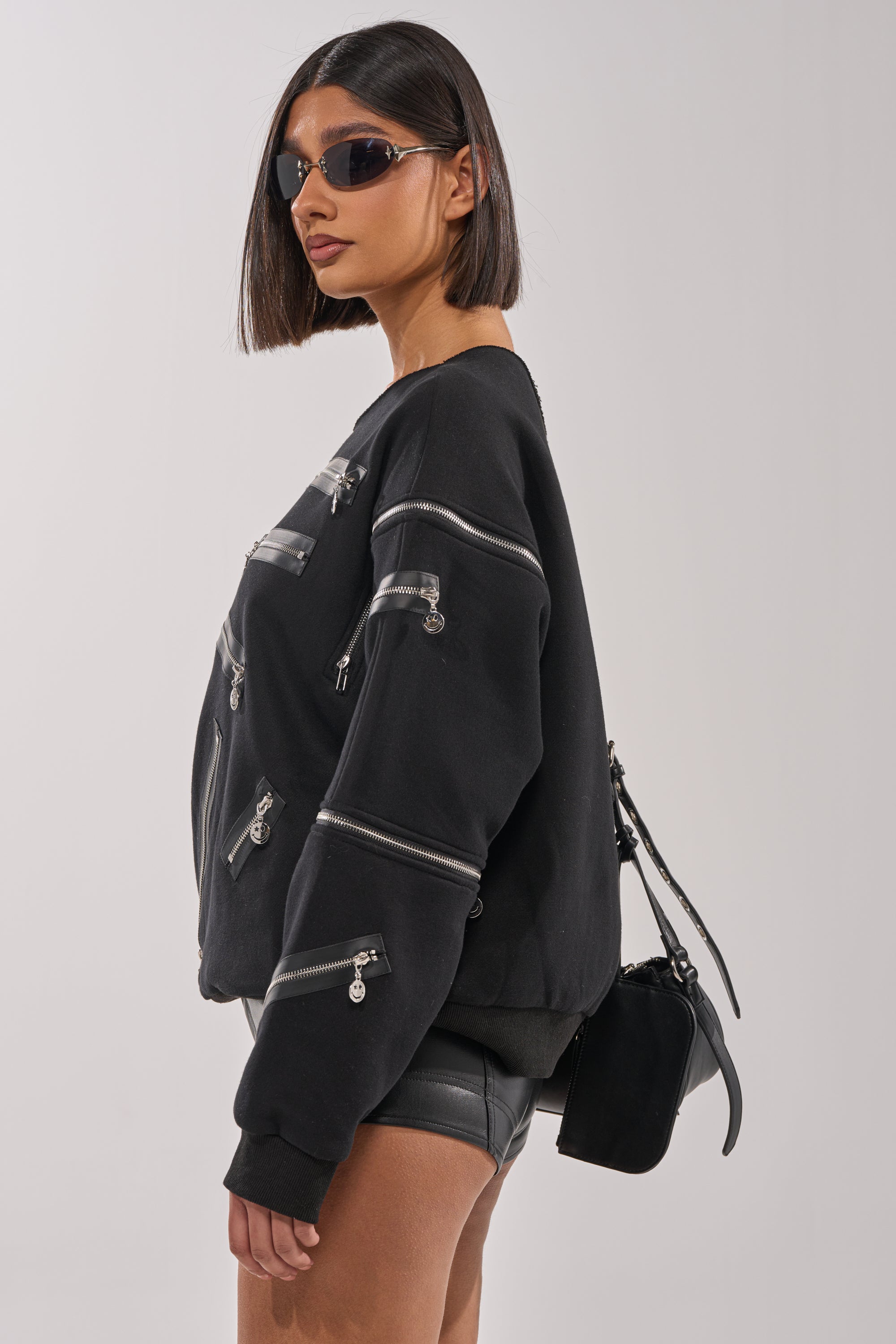 A woman with a blunt bob wears narrow sunglasses, black shorts, and carries a small black purse. She stands sideways against a plain background in the UN-ZIP ME OFF THE SHOULDER SWEATSHIRT featuring multiple silver zippers. 5