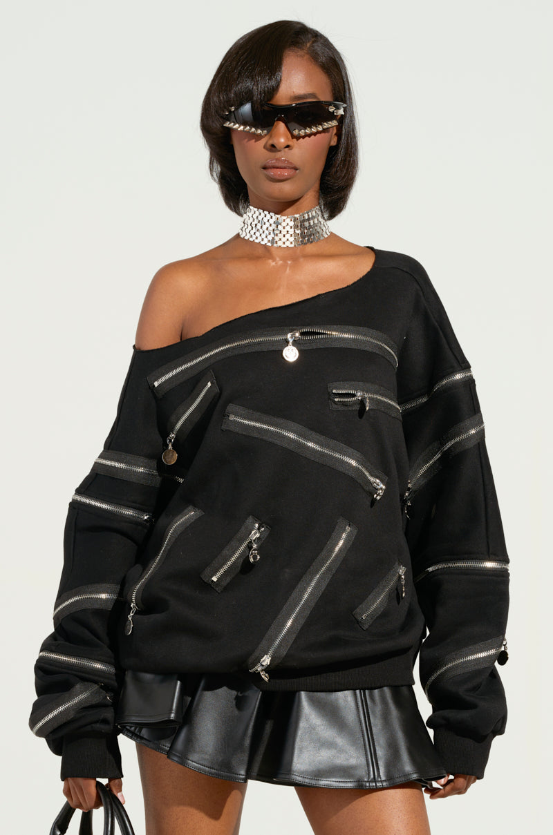 A person wearing sunglasses and a choker necklace poses confidently in the "ZIP ME UP OFF THE SHOULDER SWEATSHIRT," featuring smiley face zippers, paired with a black leather skirt. They hold a small black clutch.