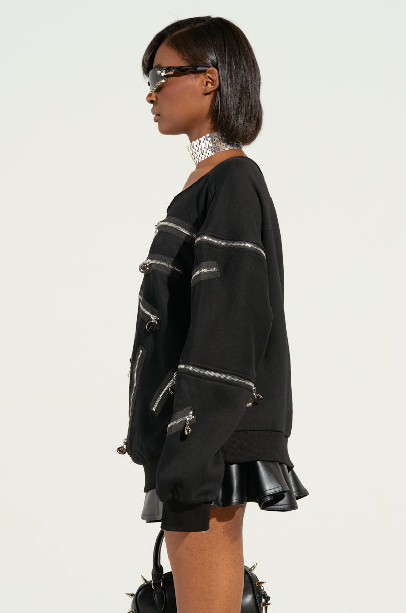 A person with short hair, wearing sunglasses and a sleek black outfit featuring smiley face zippers and a silver choker, stands in profile view. The ZIP ME UP OFF THE SHOULDER SWEATSHIRT adds flair as they hold a handbag with circular details against an off-white background.