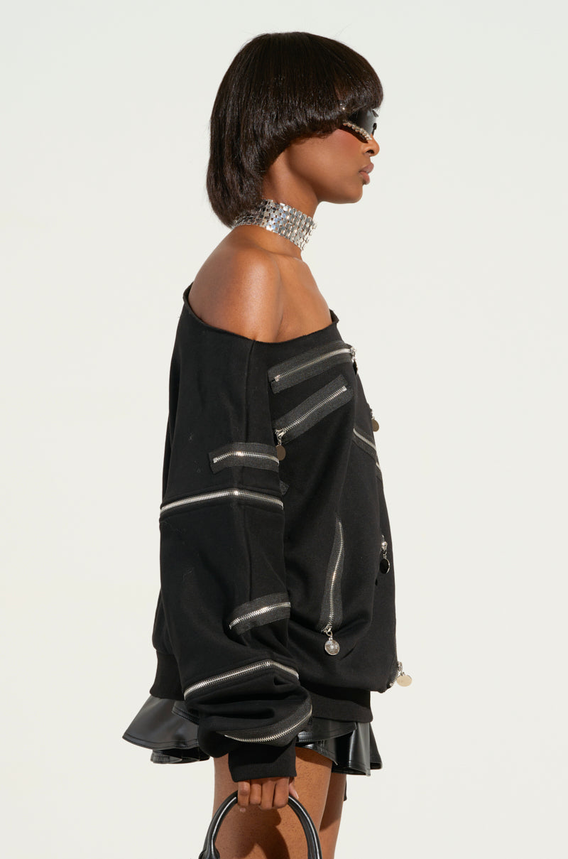 A person with a short hairstyle and sunglasses is wearing the ZIP ME UP OFF THE SHOULDER SWEATSHIRT, featuring smiley face zippers, alongside a shiny choker. They are holding a small bag and are depicted in profile against a plain background.