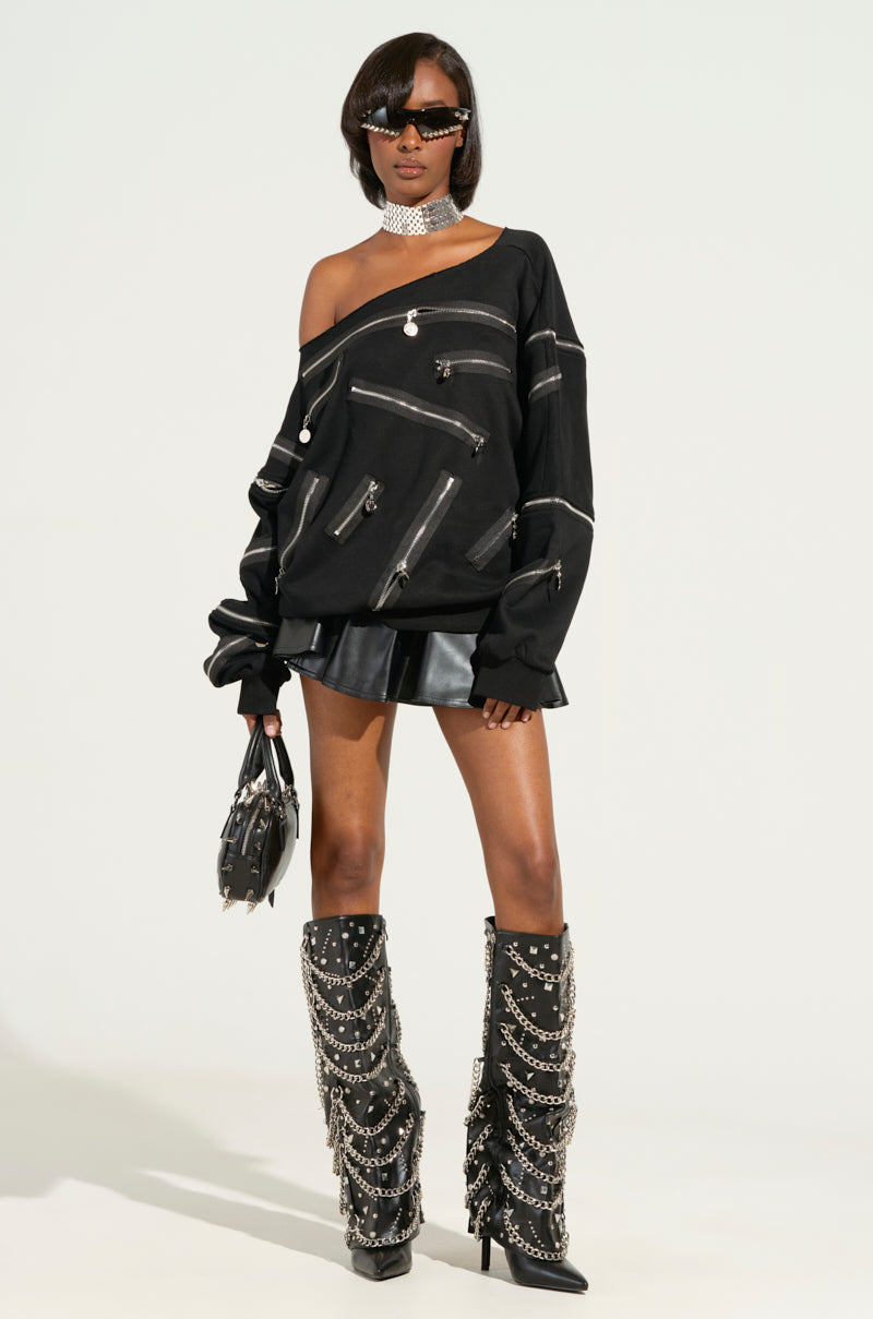 A trendy individual wears the ZIP ME UP OFF THE SHOULDER SWEATSHIRT in black, featuring smiley face zippers, paired with a leather skirt and knee-high boots adorned with chains. They accessorize with a choker and sunglasses and carry a studded handbag, posing confidently against a plain backdrop.