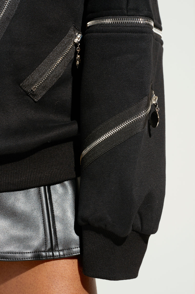 The image captures a person wearing the "ZIP ME UP OFF THE SHOULDER SWEATSHIRT," featuring smiley face zippers on the sleeves and pockets. The sweatshirt, paired with a metallic silver skirt, stands out against a light background.