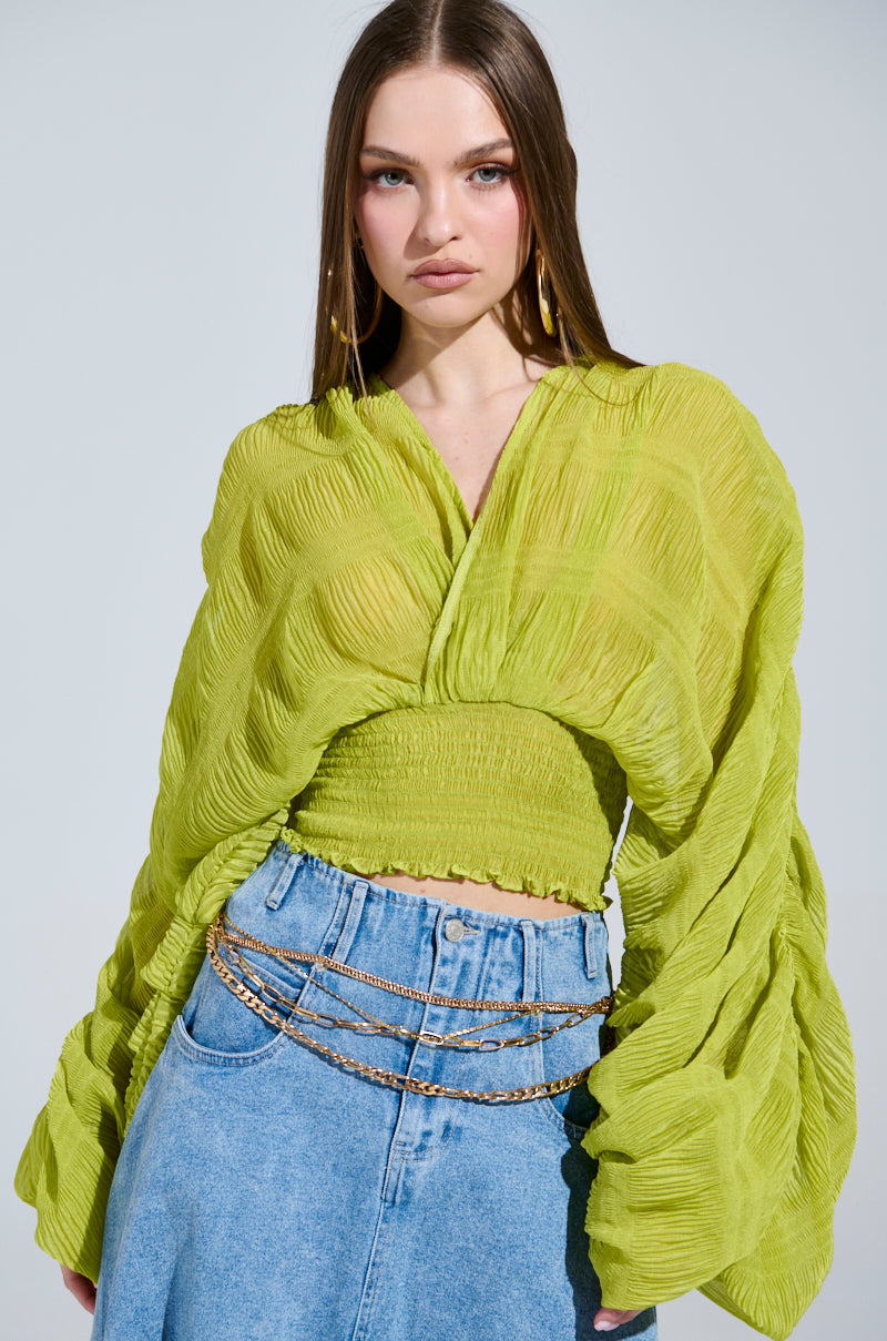 A person poses against a gray background, wearing an ALL THE BUZZ DEEP V LONG SLEEVE BLOUSE in lime green, paired with wide-leg jeans. They accessorize with layered gold chain belts and hoop earrings, exuding a calm yet confident expression.