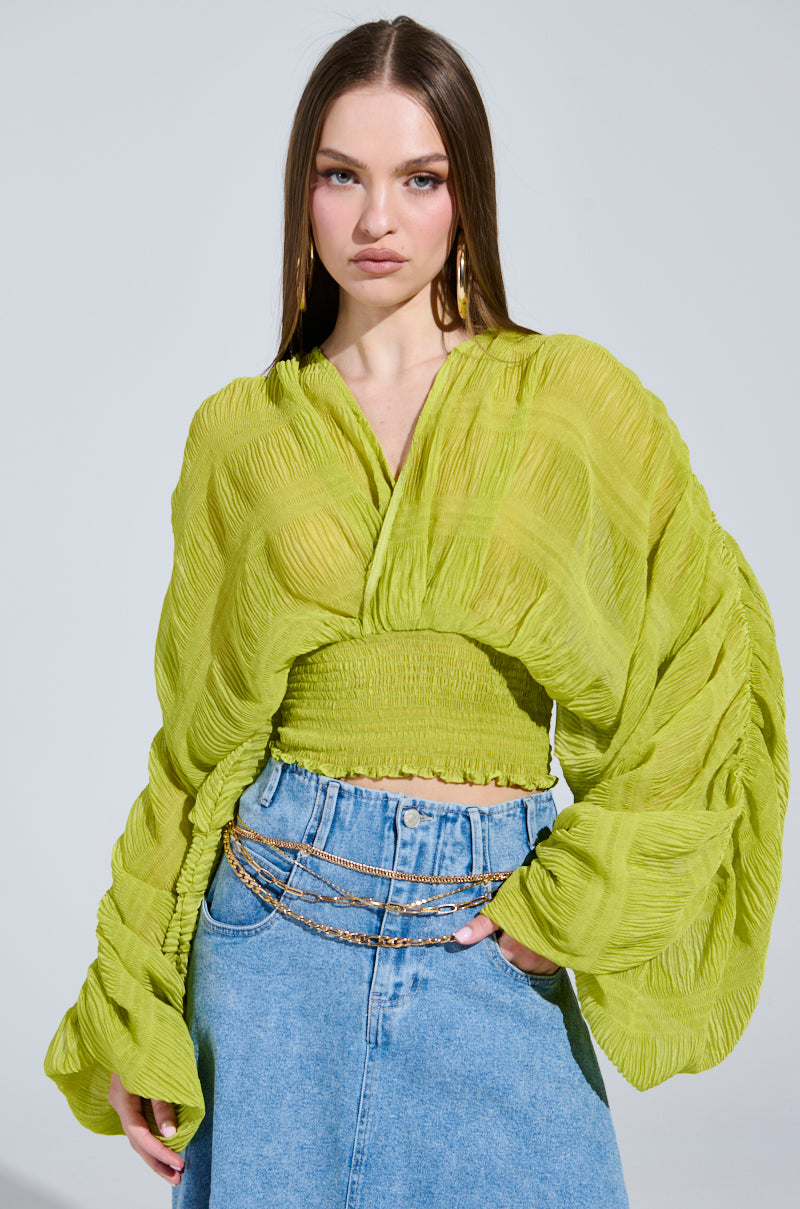 A person with long brown hair wears the ALL THE BUZZ DEEP V LONG SLEEVE BLOUSE in bright green, featuring oversized sleeves and a smocked waist, paired with blue high-waisted denim jeans. They accessorize with a layered chain belt and large hoop earrings against a light background.