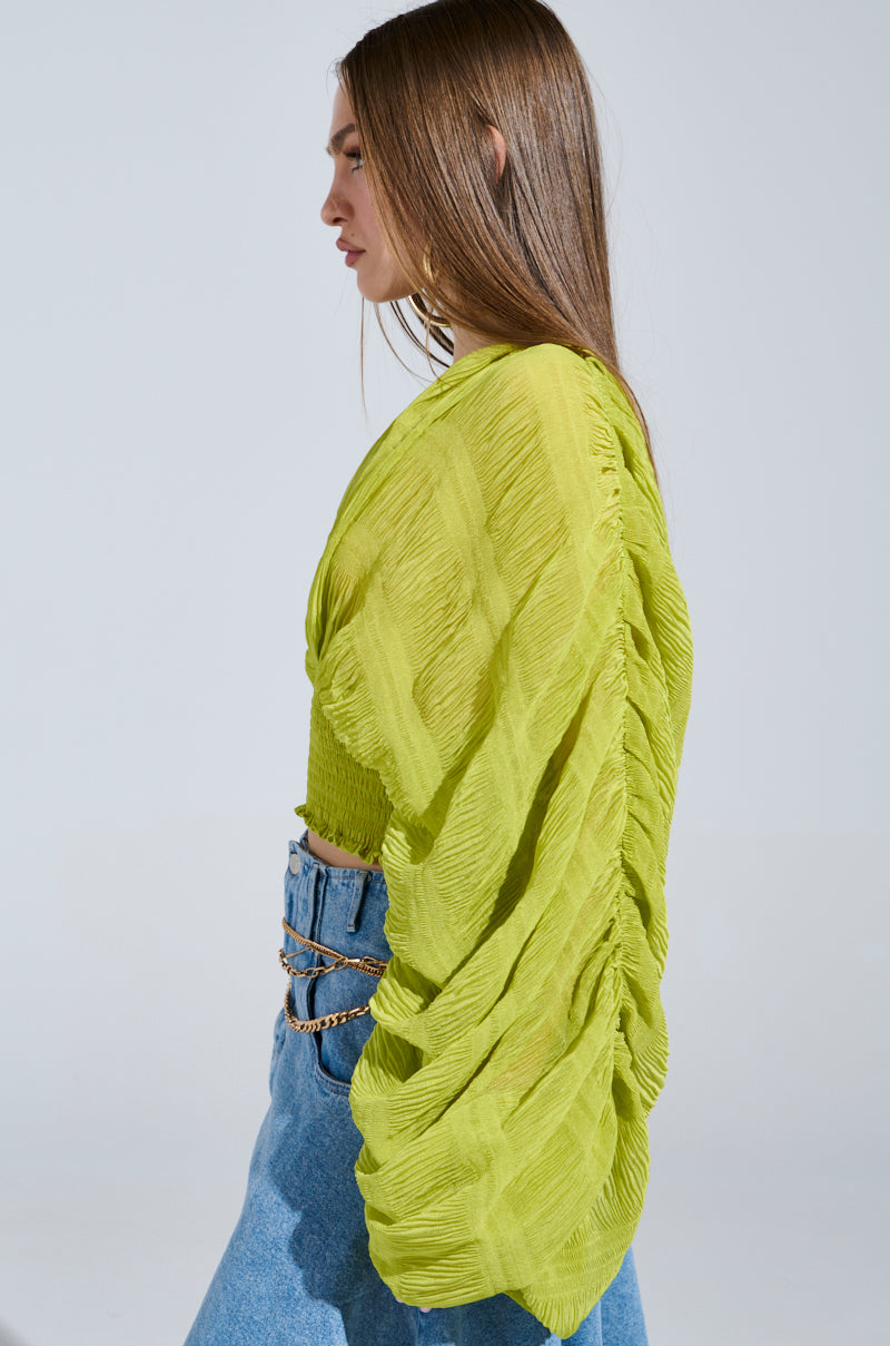 A woman stands in profile, donning the ALL THE BUZZ DEEP V LONG SLEEVE BLOUSE in a bright green, textured draped design with an oversized fit. She pairs it with blue jeans accentuated by a chain belt. Her sleek, straight brown hair adds to the clean look against the plain background.