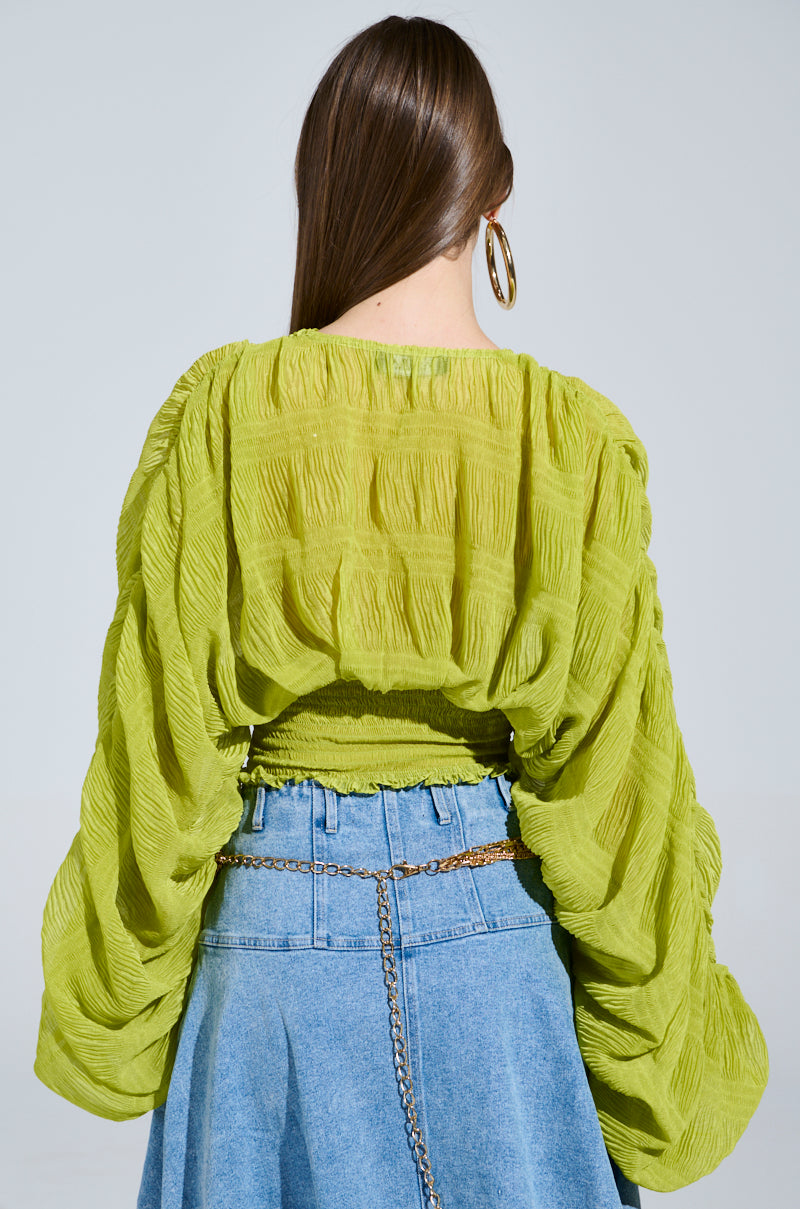A woman with long brown hair is styled from behind in a vibrant ALL THE BUZZ DEEP V LONG SLEEVE BLOUSE featuring puffy, semi-sheer sleeves, paired with a denim skirt accented by a gold chain belt and large hoop earrings. The backdrop is a soft gray.