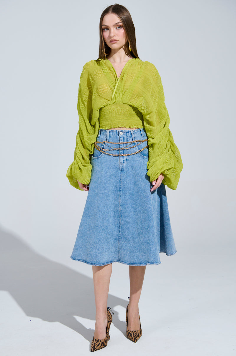 A woman sports the ALL THE BUZZ DEEP V LONG SLEEVE BLOUSE in textured lime green, paired with a blue denim skirt highlighted by a gold chain belt. She completes the look with animal print pointed-toe heels against a plain light gray background.