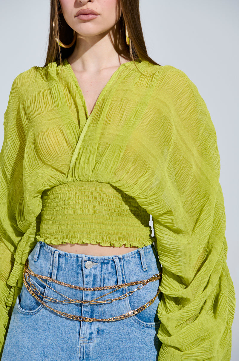 A woman wears the "ALL THE BUZZ DEEP V LONG SLEEVE BLOUSE," a semi-sheer lime green top with wide sleeves and a smocked waist. She pairs it with hoop earrings, layered gold chains, and blue jeans, adding effortless chic to the soft gray backdrop.