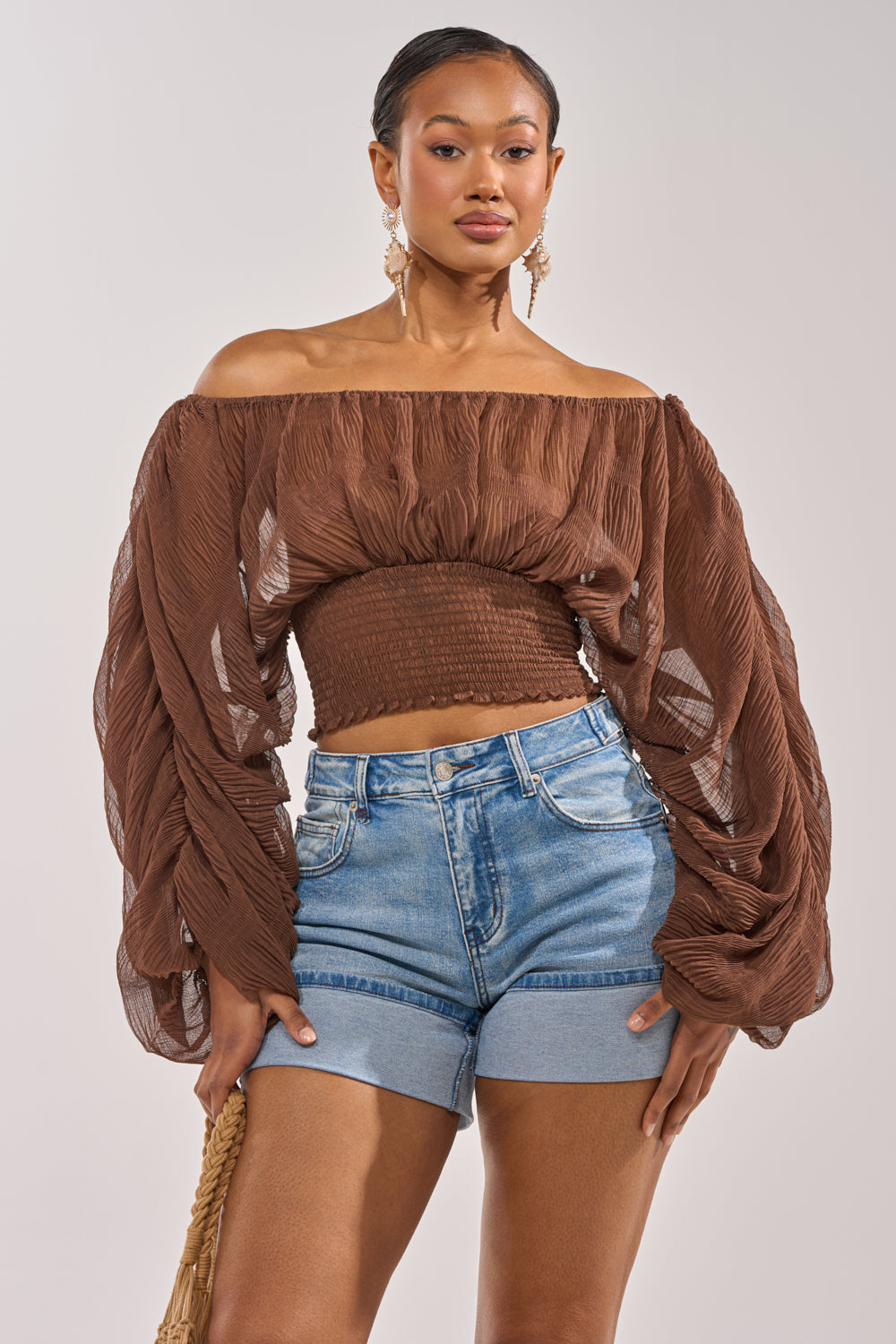 A woman poses confidently in the DIVINE RIGHT LONG SLEEVE BLOUSE IN BROWN, an off-the-shoulder sheer top with romantic volume, paired with high-waisted denim shorts, statement earrings, and a woven bag for a chic daytime look. 0