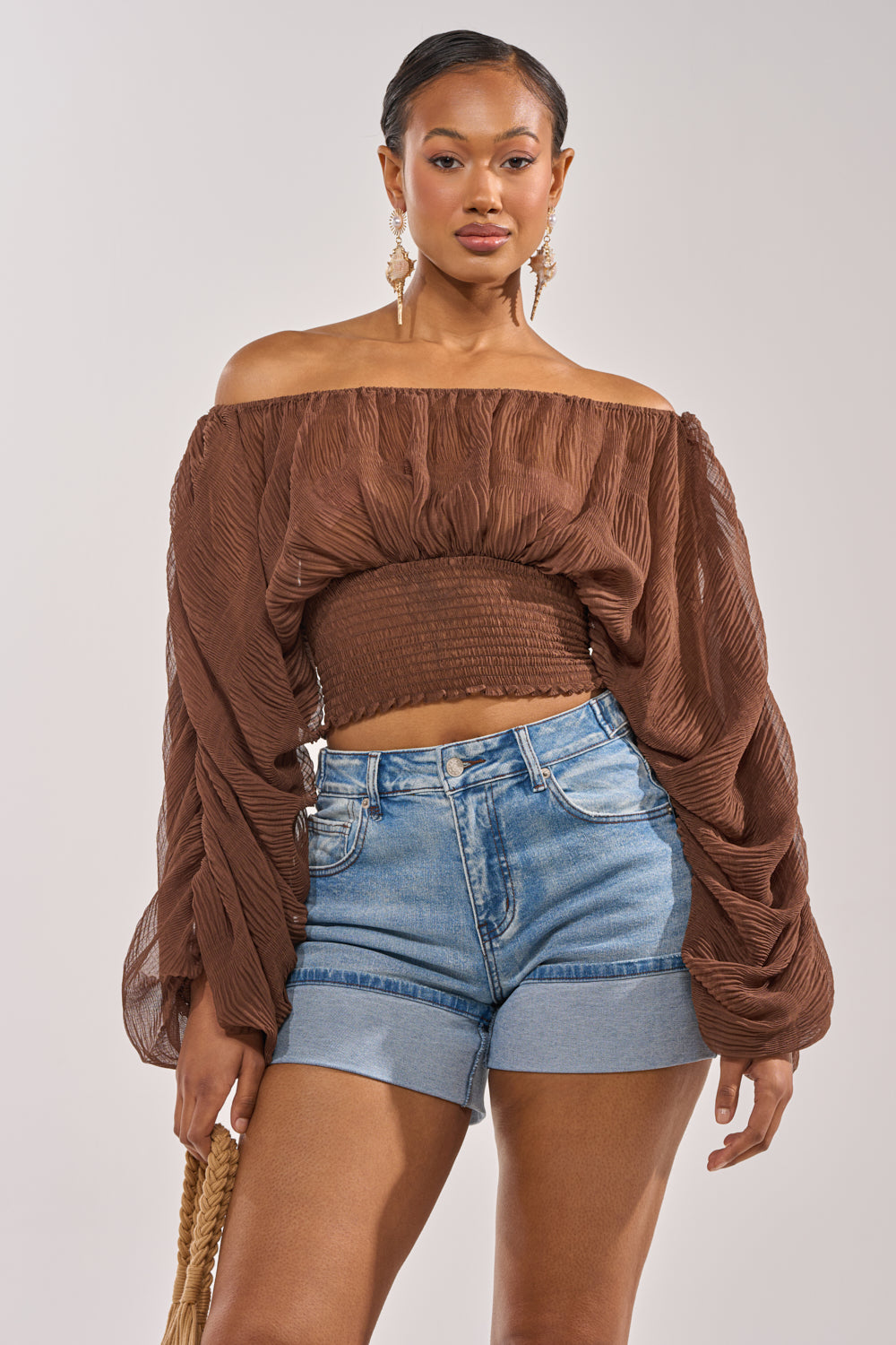 Wearing the DIVINE RIGHT LONG SLEEVE BLOUSE IN BROWN, a woman faces forward in high-waisted rolled-hem denim shorts, holding a woven bag. Her hair is pulled back, and she accessorizes with gold earrings. 1