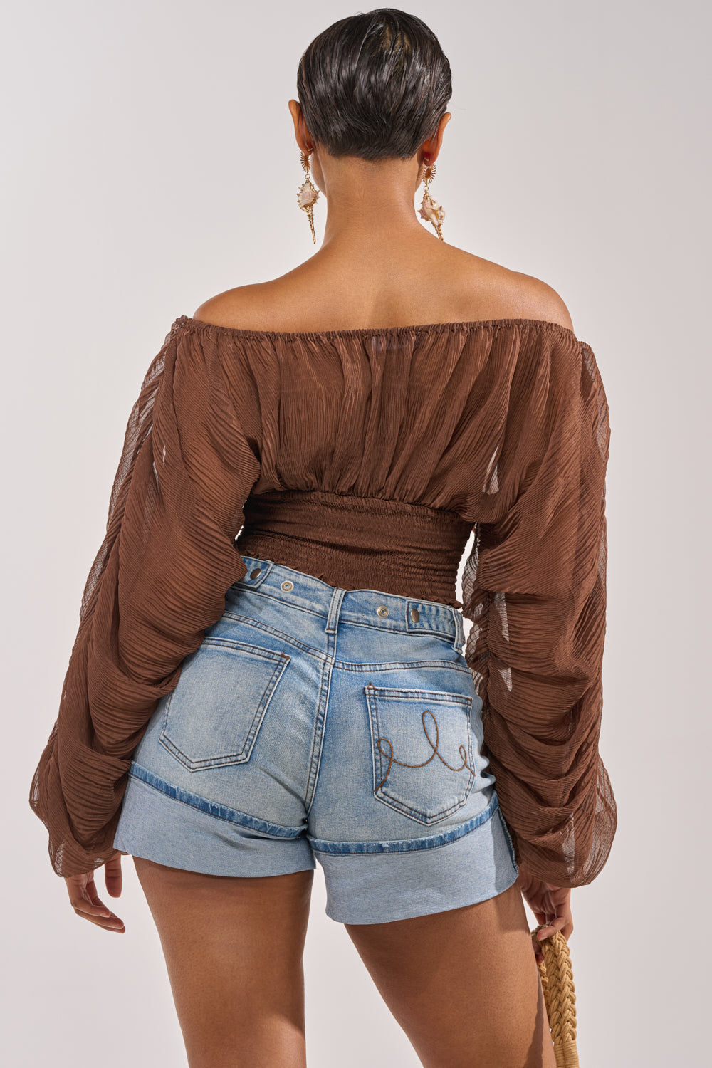 A woman with short hair is seen from behind wearing the DIVINE RIGHT LONG SLEEVE BLOUSE IN BROWN and light blue high-waisted denim shorts with unique pocket stitching, holding a woven bag in her left hand. 3