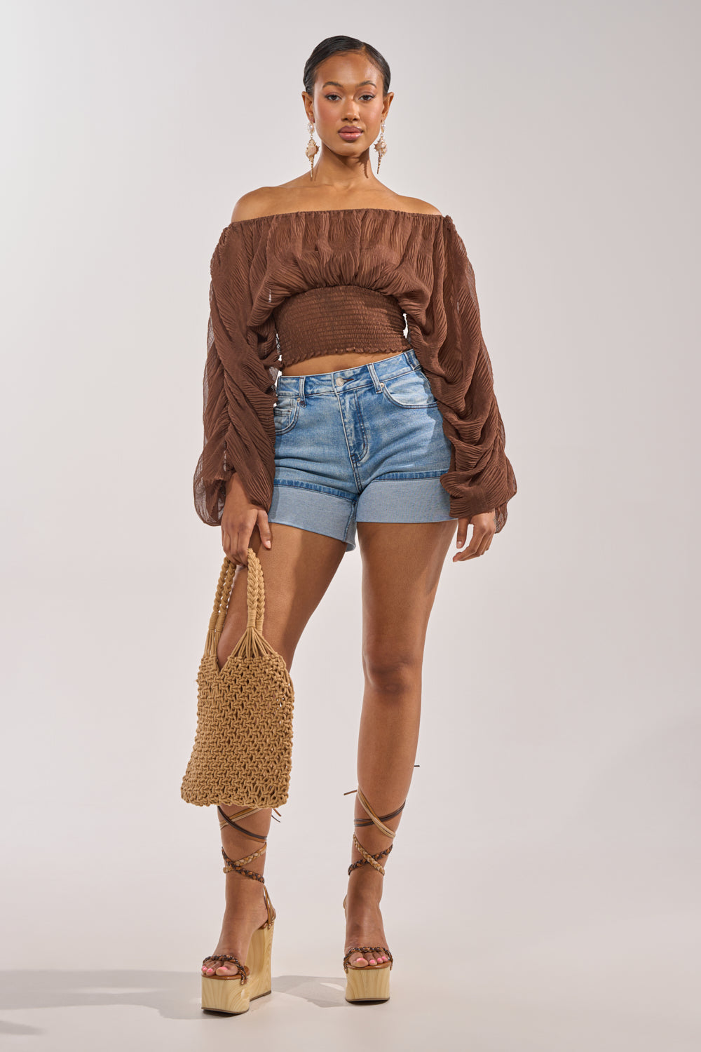 A woman stands confidently wearing the DIVINE RIGHT LONG SLEEVE BLOUSE IN BROWN, paired with blue denim shorts, platform lace-up sandals, a woven straw bag, and hoop earrings. 4