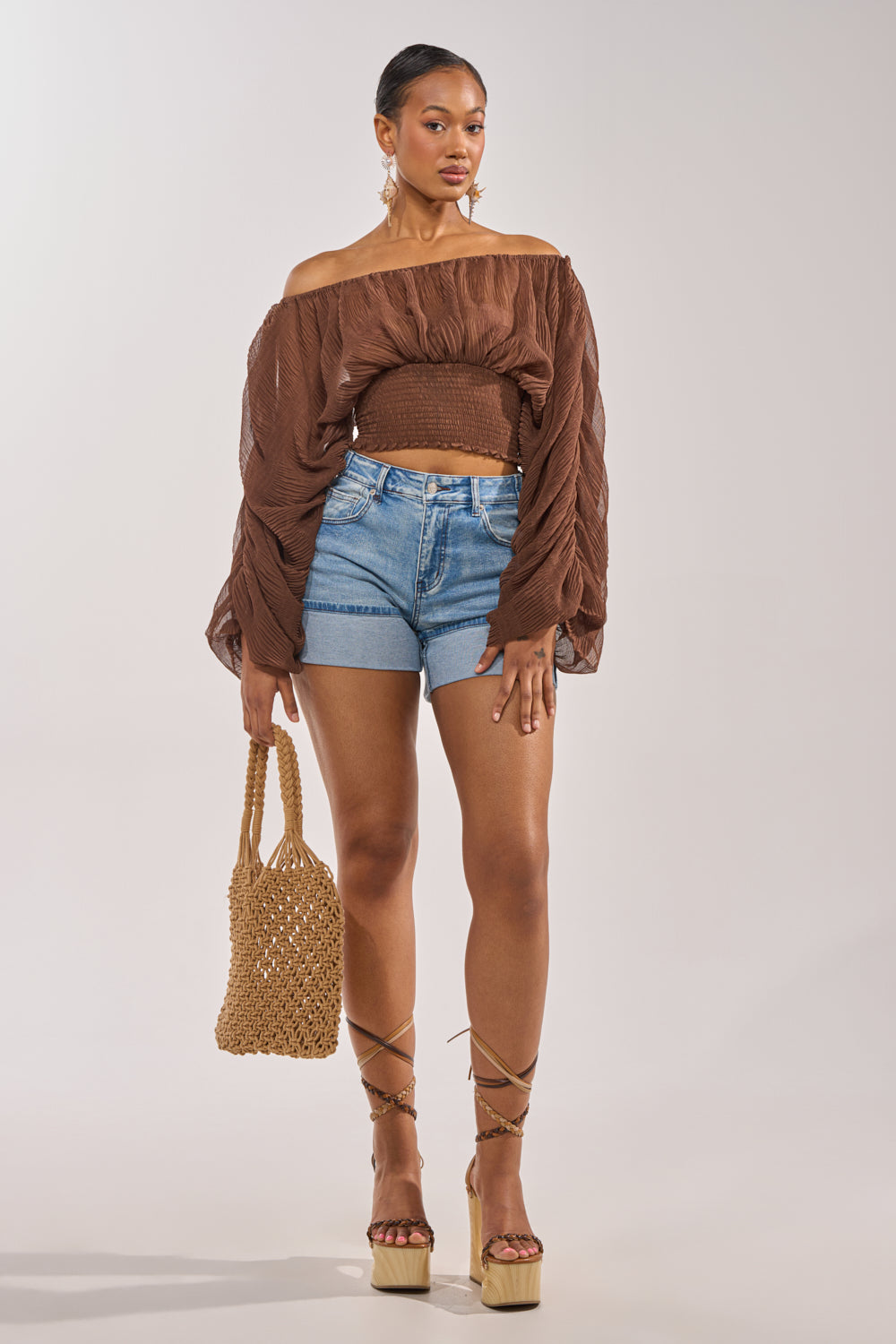A woman poses against a plain light background, wearing the DIVINE RIGHT LONG SLEEVE BLOUSE IN BROWN with denim shorts and platform sandals, holding a woven bag. 5