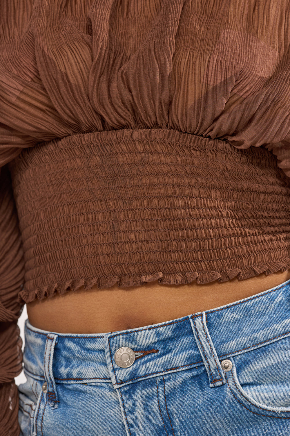 A close-up of a person wearing the DIVINE RIGHT LONG SLEEVE BLOUSE IN BROWN, featuring textured fabric and a smocked waist, paired with high-waisted light blue denim jeans. The focus is on the gathered midsection and elastic waistband of the top. 6