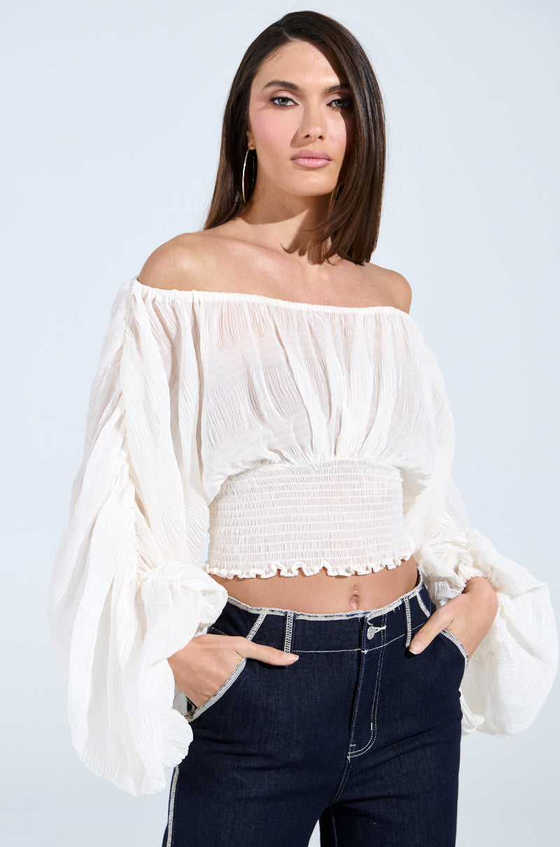 A woman stands hands-in-pockets against a plain background, wearing the KOA Flowy Pleated Long Sleeve Blouse in white with smocked details and off-the-shoulder design, paired with dark high-waisted jeans.