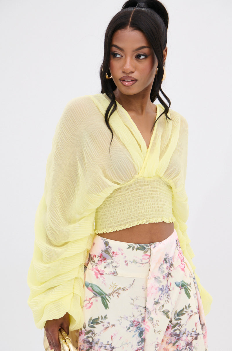 A person in a flowing DIVINE RIGHT LONG SLEEVE BLOUSE TOP and a pastel floral skirt looks aside. The lemon yellow chiffon blouse features sheer sleeves and a textured pattern, while their long dark hair is tied back against a plain backdrop.