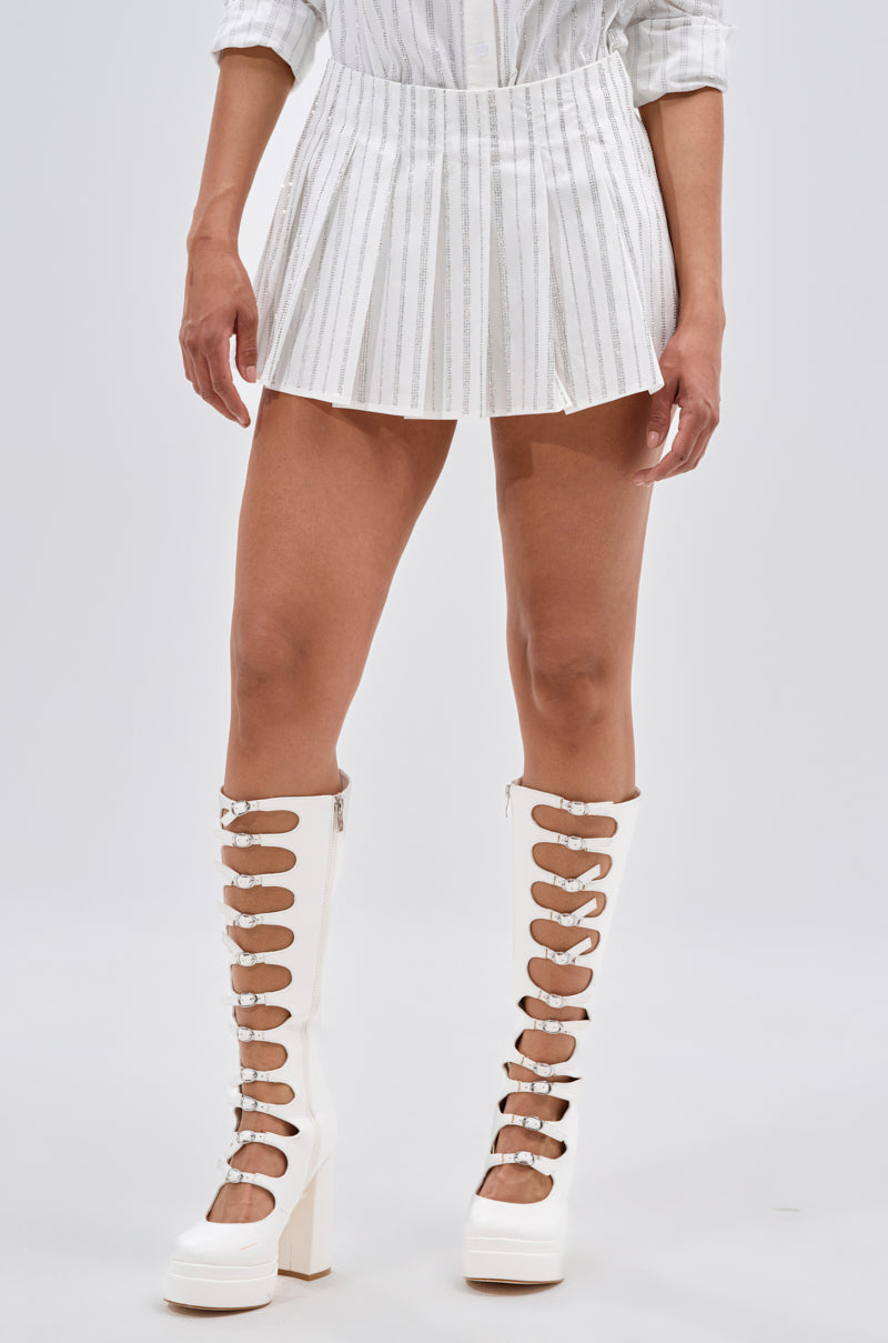 Wearing the ONTO THE NEXT EMBELLISHED PLEATED MINI SKIRT IN WHITE and knee-high white platform boots with multiple buckled straps, a person stands against a plain light background. 1