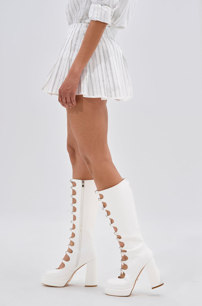 A person wears the ONTO THE NEXT EMBELLISHED PLEATED MINI SKIRT IN WHITE with white knee-high platform boots featuring circular cutouts and lace-up fronts, standing against a plain light background. 3