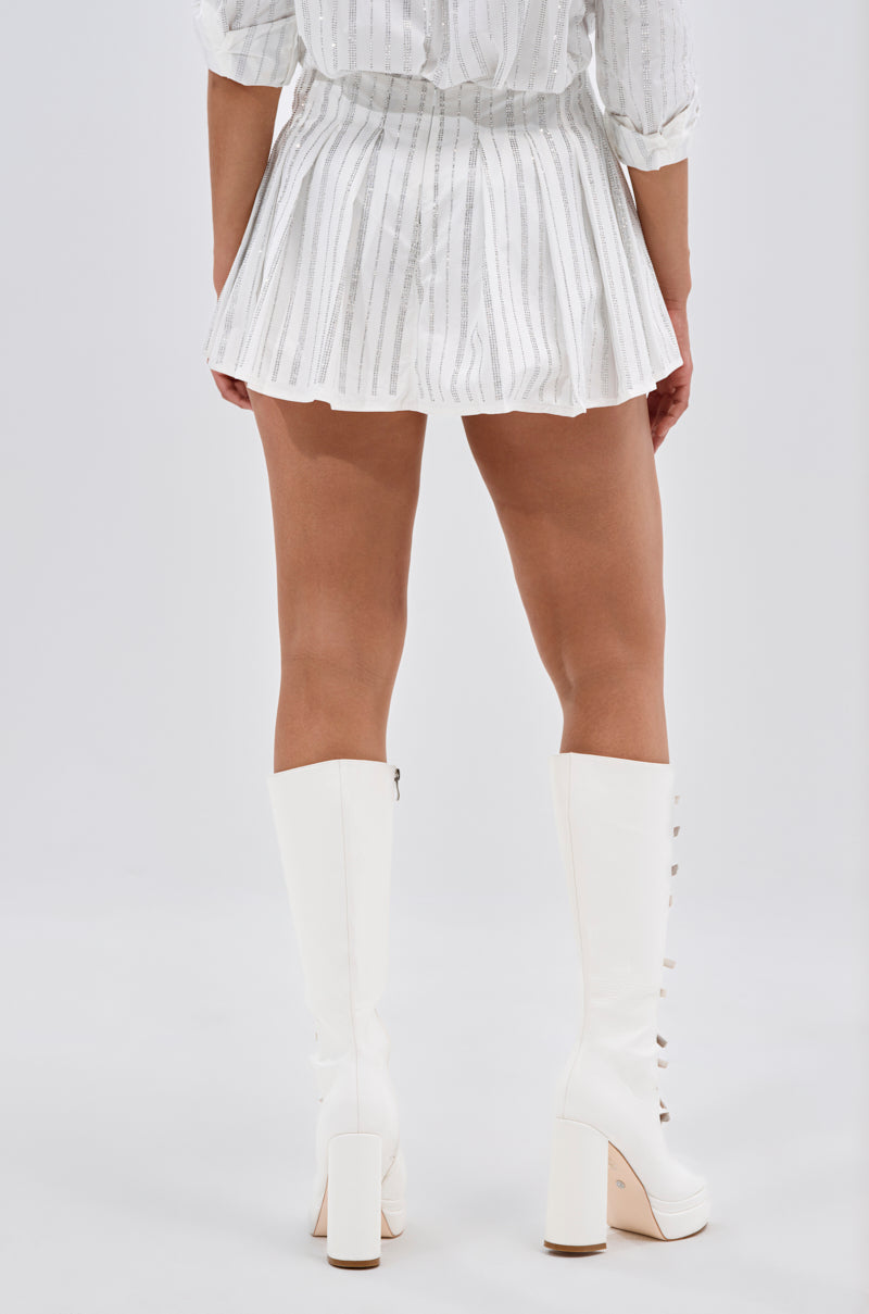 A person faces away from the camera, modeling the ONTO THE NEXT EMBELLISHED PLEATED MINI SKIRT IN WHITE with a white long-sleeve top and tall white heeled boots against a plain backdrop. 4