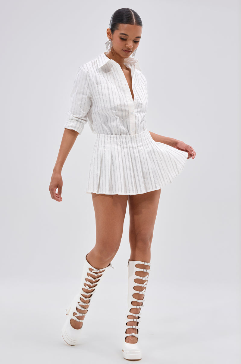 A woman wears the ONTO THE NEXT EMBELLISHED PLEATED MINI SKIRT IN WHITE with a white, pinstriped collared shirt dress and knee-high lace-up boots, walking on a light background while looking down and holding her skirt. 5