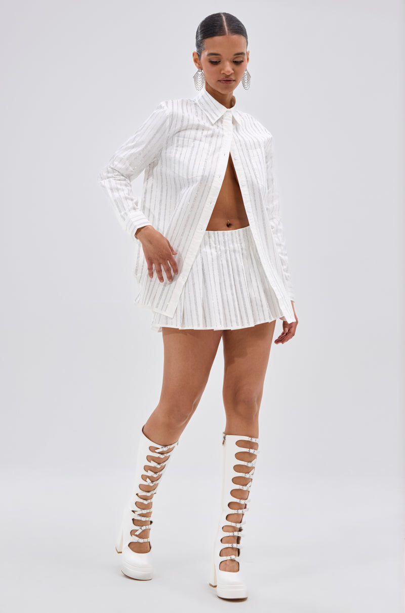 A woman stands against a plain light background wearing the ONTO THE NEXT EMBELLISHED PLEATED MINI SKIRT IN WHITE, a long-sleeve pinstriped shirt, statement earrings, and knee-high white lace-up boots. 6