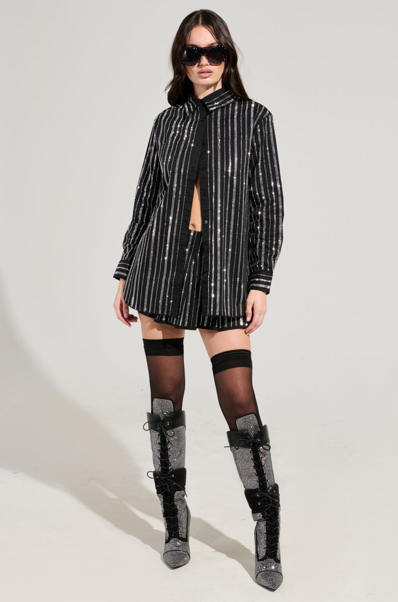 A woman in sunglasses wears a black button-up shirt and a pleated mini skirt, paired with rhinestone-embellished over-the-knee black socks and sparkly silver and black lace-up boots. She stands confidently against a plain light background.