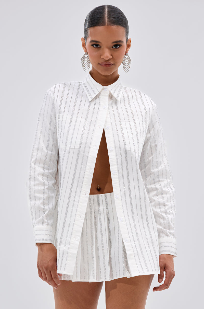 A woman with slicked-back hair and statement earrings wears the ONTO THE NEXT EMBELLISHED BUTTON DOWN TOP IN WHITE, featuring vertical stripes, paired with a matching high-waisted mini skirt against a plain light gray background. 0