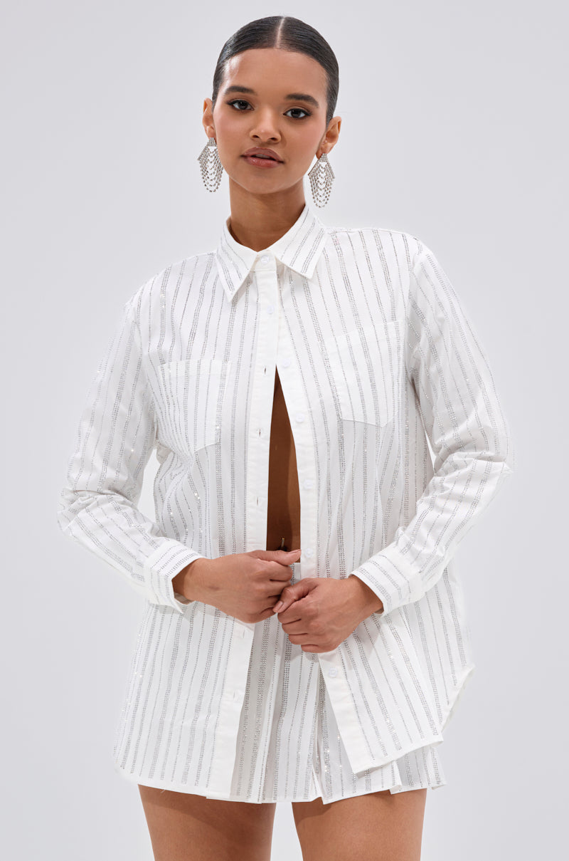 A woman wears the ONTO THE NEXT EMBELLISHED BUTTON DOWN TOP IN WHITE, featuring long sleeves, a cotton blend fabric, and silver vertical stripes. She pairs it with matching shorts and stands against a plain light gray background. 1