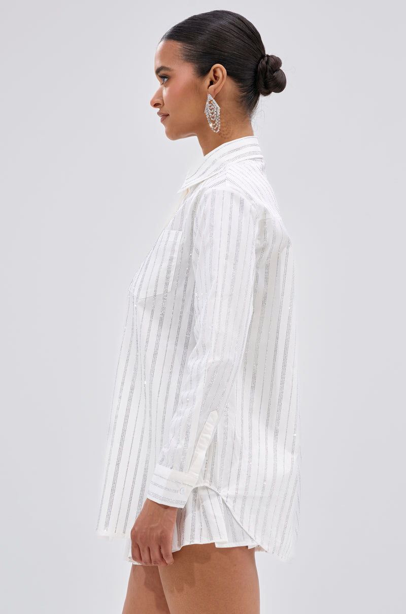 A woman with dark hair in a low bun stands in profile against a light gray background, wearing the ONTO THE NEXT EMBELLISHED BUTTON DOWN TOP IN WHITE with long sleeves, vertical thin black stripes, and dangling earrings. 2