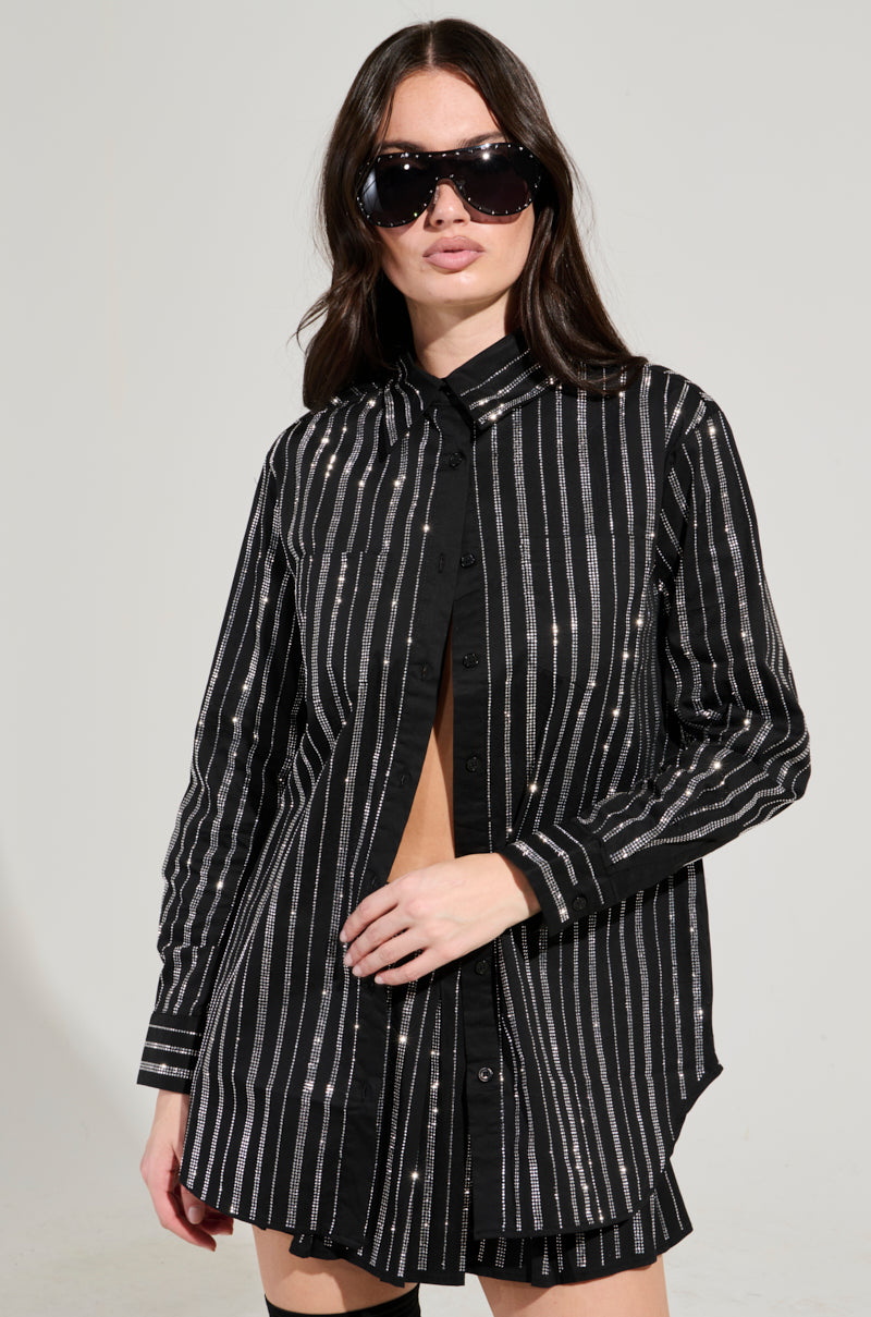 A woman in oversized sunglasses confidently models the ONTO THE NEXT EMBELLISHED BUTTON DOWN TOP against a plain background. This black shirt features vertical white stripes and rhinestone embellishments, which she holds slightly open, while her long hair cascades freely for a chic and fashionable look.