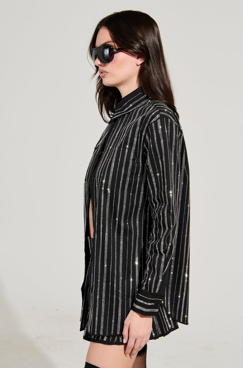 A person with long dark hair wears black sunglasses and the ONTO THE NEXT EMBELLISHED BUTTON DOWN TOP, featuring subtle rhinestone decorations. They stand against a plain background, looking to the side.