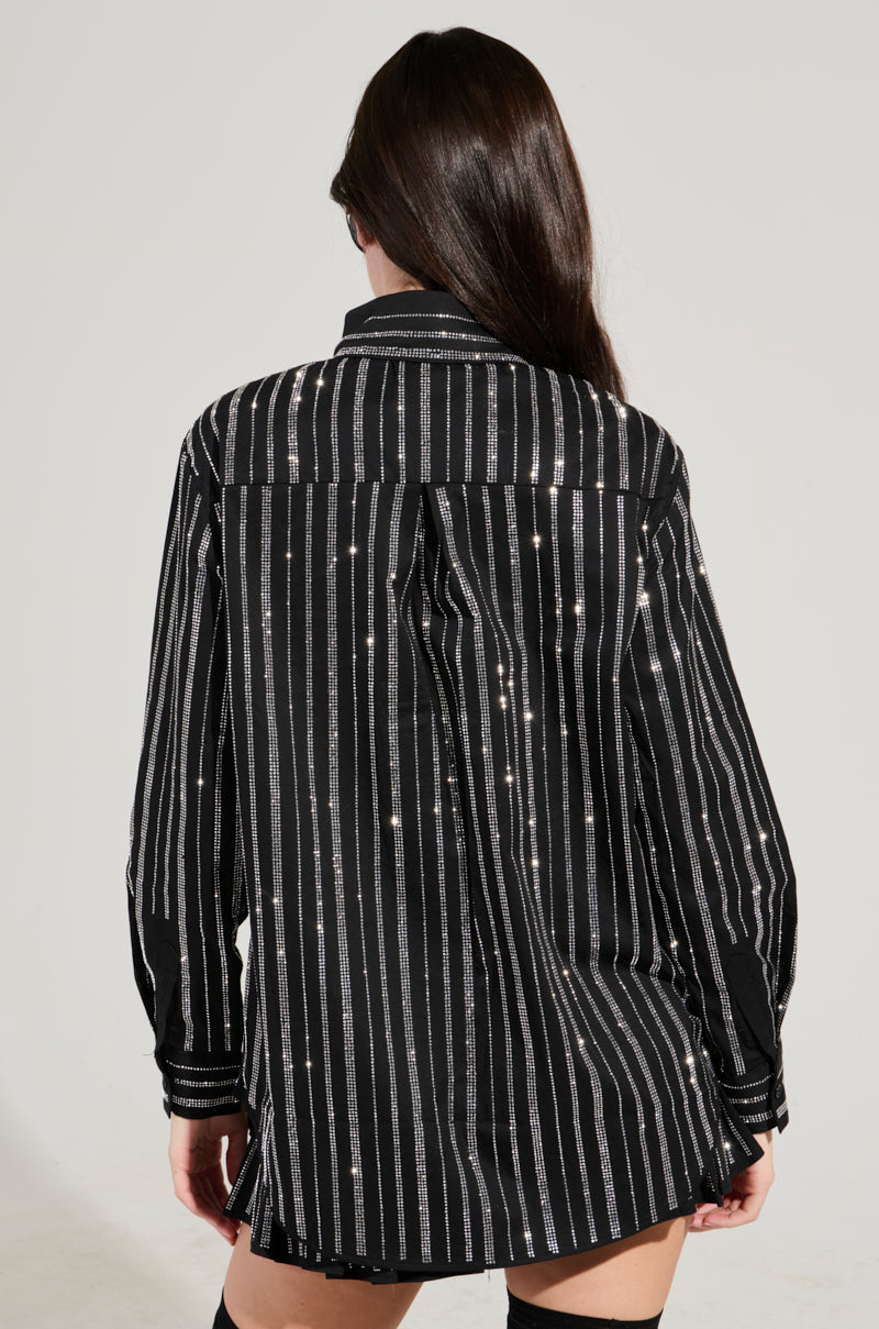 A person with long dark hair faces away from the camera, wearing the ONTO THE NEXT EMBELLISHED BUTTON DOWN TOP. This oversized black shirt features vertical silver stripes, small embroidered stars, and subtle rhinestone embellishments that add a touch of sparkle to the outfit. They complete the look with sleek black thigh-high socks.