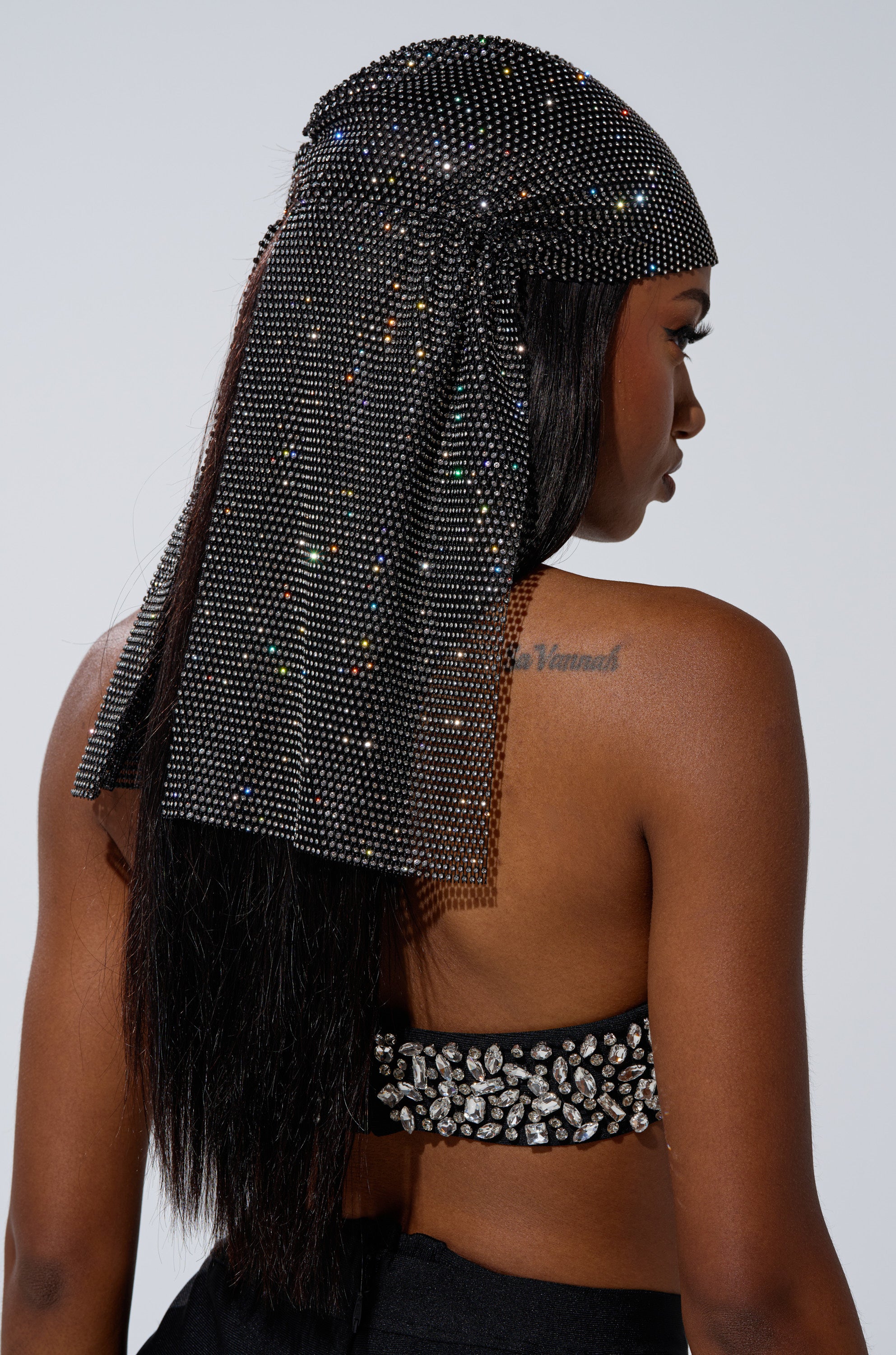 A woman with long, straight hair and a tattoo on her right shoulder blade wears the ELEGANCE IS EARNED HEADPIECE, featuring sparkling rhinestones and an elasticized backing, paired with a bejeweled black top.