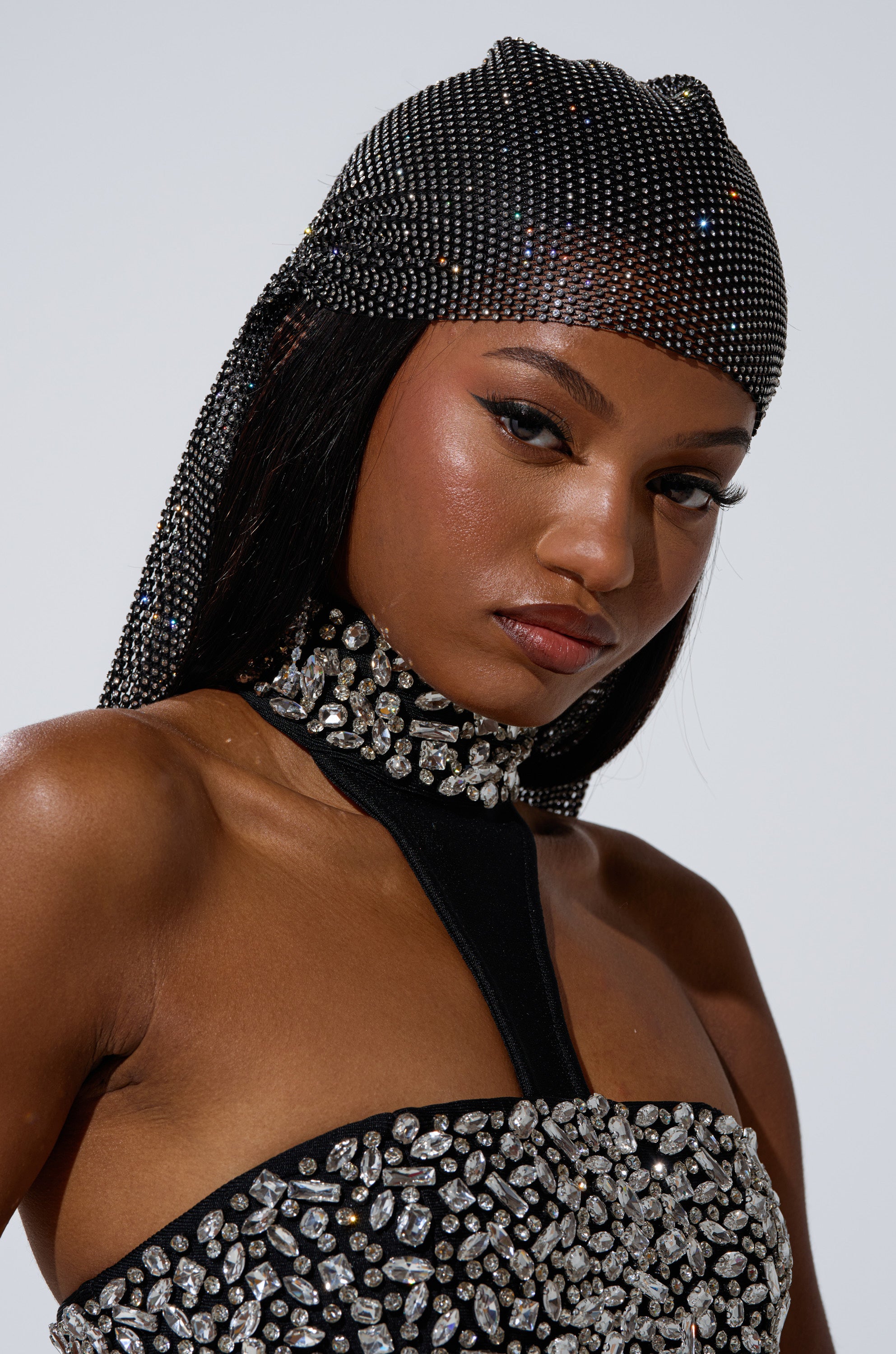 A woman wears the ELEGANCE IS EARNED HEADPIECE, a black sparkling rhinestone accessory, paired with a matching choker and top. She looks confident, her glowing skin and mesh hair accents standing out against a plain background.