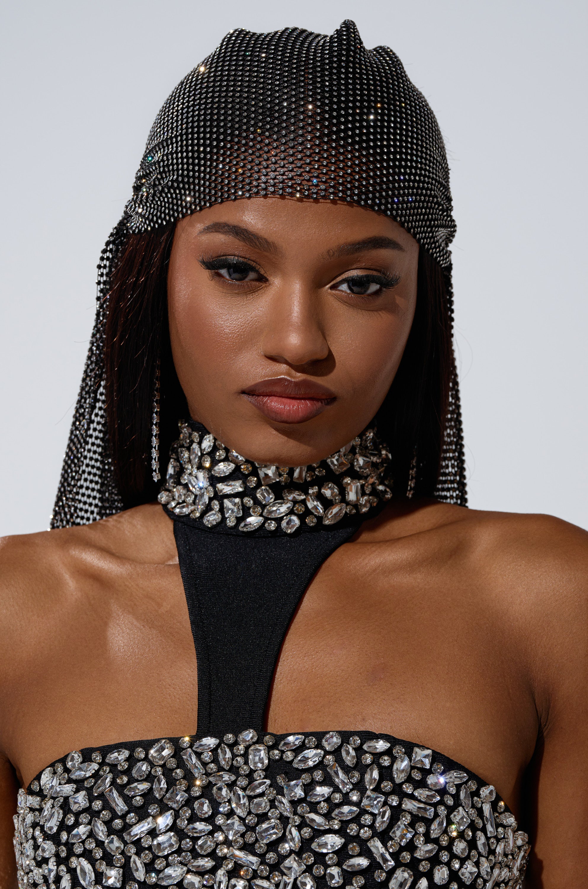 A woman wearing a black outfit with sparkling rhinestones and the ELEGANCE IS EARNED HEADPIECE gazes confidently at the camera against a plain background.