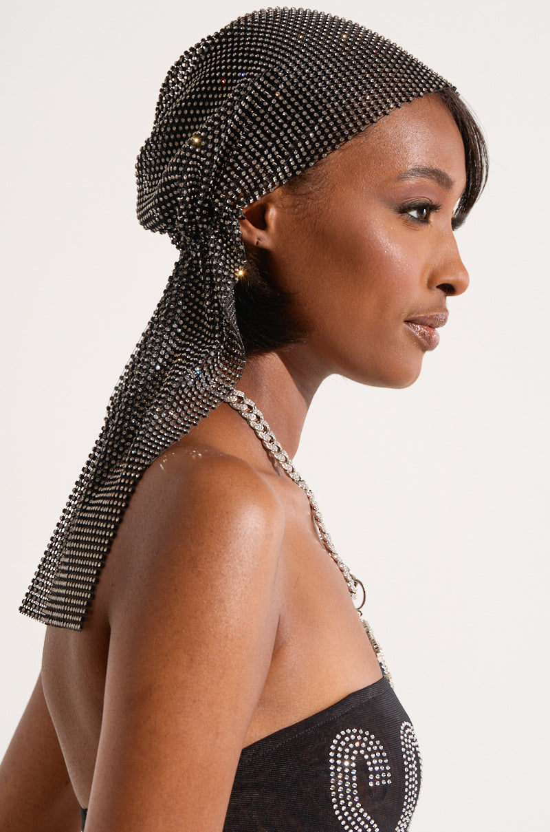 A woman is shown in profile, wearing the ELEGANCE IS EARNED HEADPIECE—a sparkly black accessory adorned with small gems. She has short hair beneath the headpiece and is dressed in a strapless black top featuring a decorative design, complemented by a chunky silver necklace. The background is plain and light-colored. The headpiece boasts an elasticized backing for a secure fit.