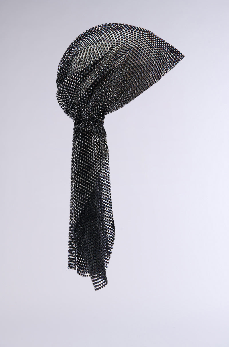 The "ELEGANCE IS EARNED HEADPIECE" is a floating black mesh scarf with a metallic sheen, artfully arranged to create an elegant, flowing shape. This versatile headpiece is photographed against a plain, light grey background, highlighting its texture and delicate pattern, showcasing its potential to double as a chic headpiece. 7