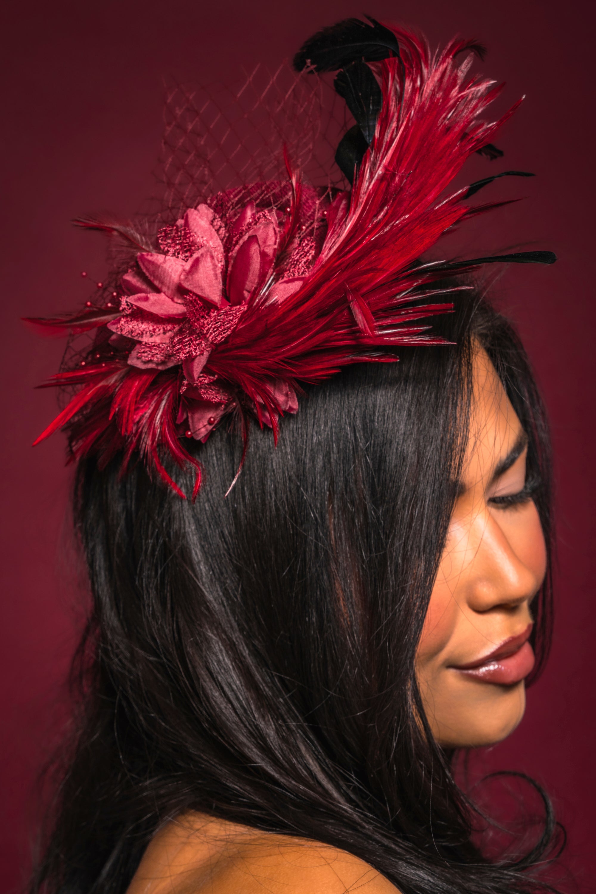 A woman with long dark hair wears the TOP OF THE DAY CLIP ON FASCINATOR, a vibrant red accessory adorned with feathers, flowers, and netting. Her eyes are closed against a matching red backdrop—an elegant choice for weddings. 0