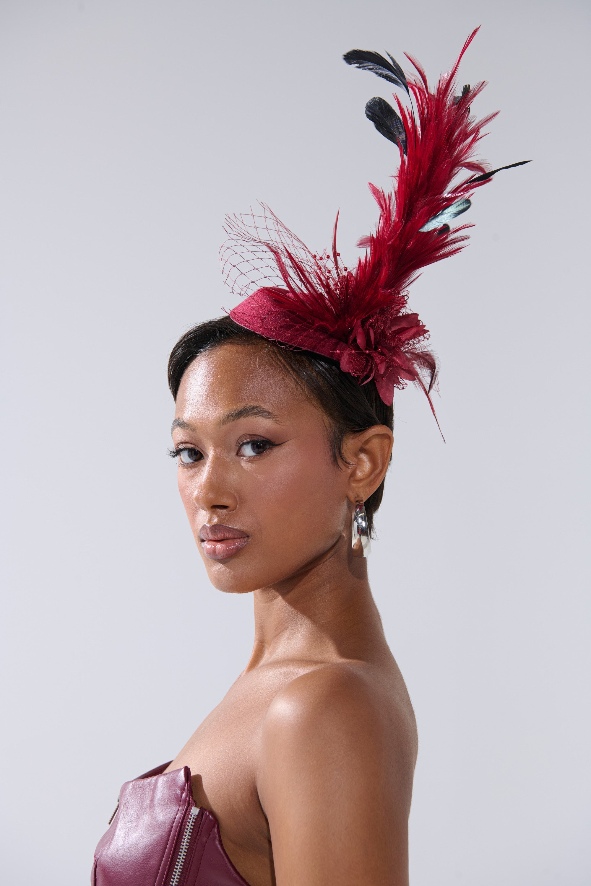 A woman in a strapless maroon dress and silver hoop earrings poses in profile, showcasing the TOP OF THE DAY CLIP ON FASCINATOR adorned with red and black feathers and netting. 2