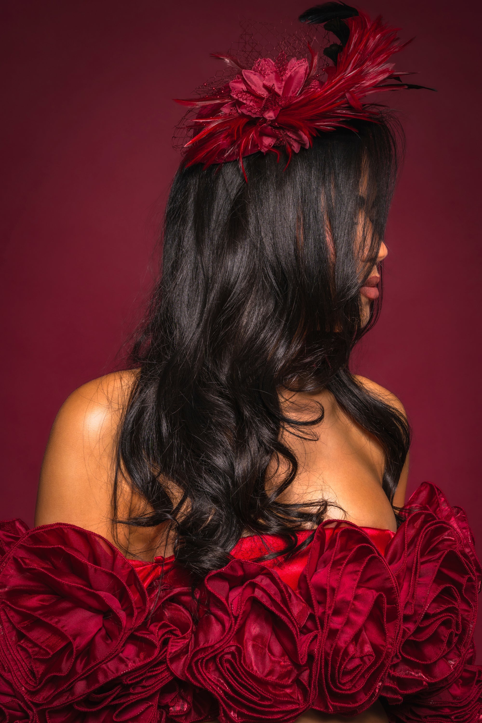 A woman with long, wavy black hair models the TOP OF THE DAY CLIP ON FASCINATOR and an off-shoulder red dress with textured floral fabric, set against a deep red background. She turns to the side, partially hiding her face. 1