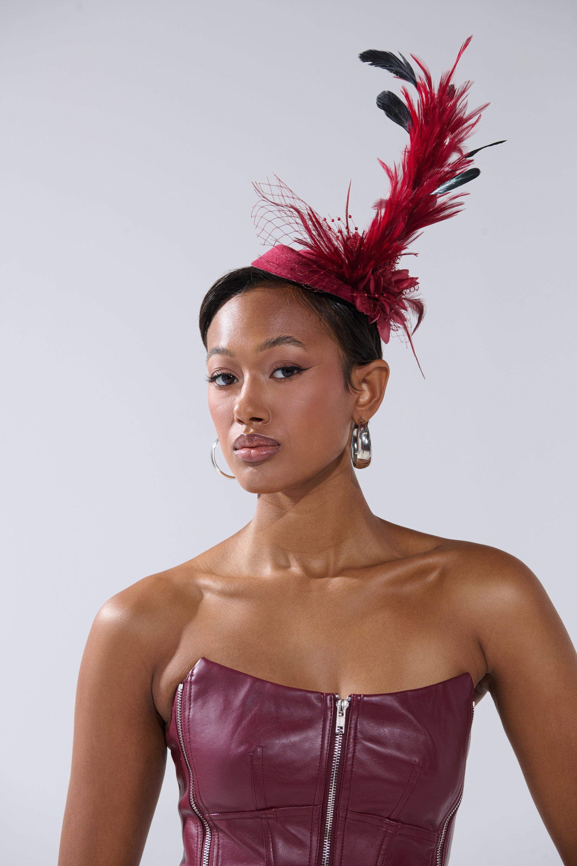 A woman poses against a plain light background wearing large hoop earrings, a strapless maroon leather dress with a front zipper, and the TOP OF THE DAY CLIP ON FASCINATOR in striking red. 3