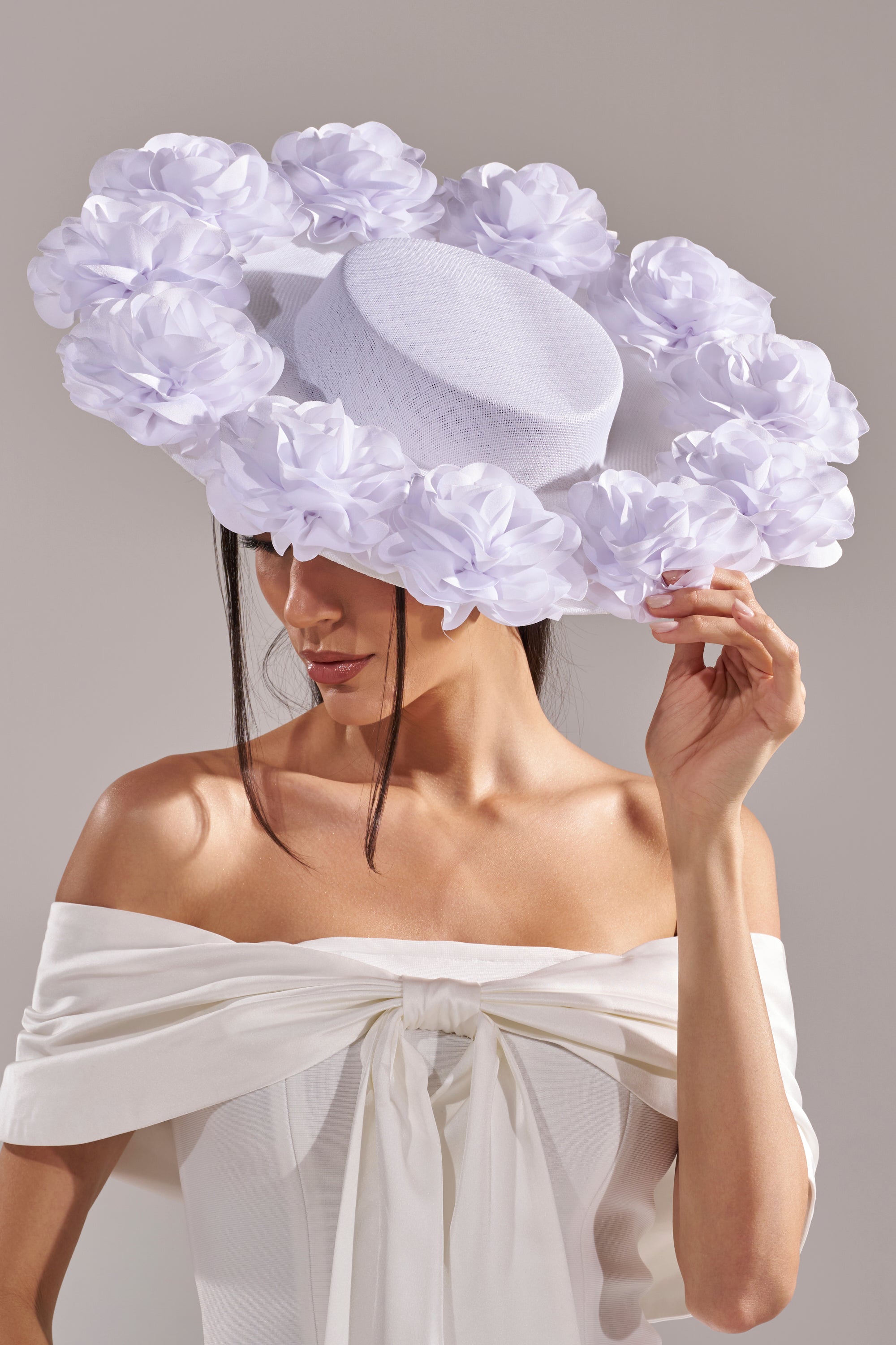 A woman in an off-the-shoulder white dress holds the brim of the ROSEY CLIP ON HAT IN WHITE, featuring oversized 3D rose detailing. The hat partially obscures her face and dark hair.