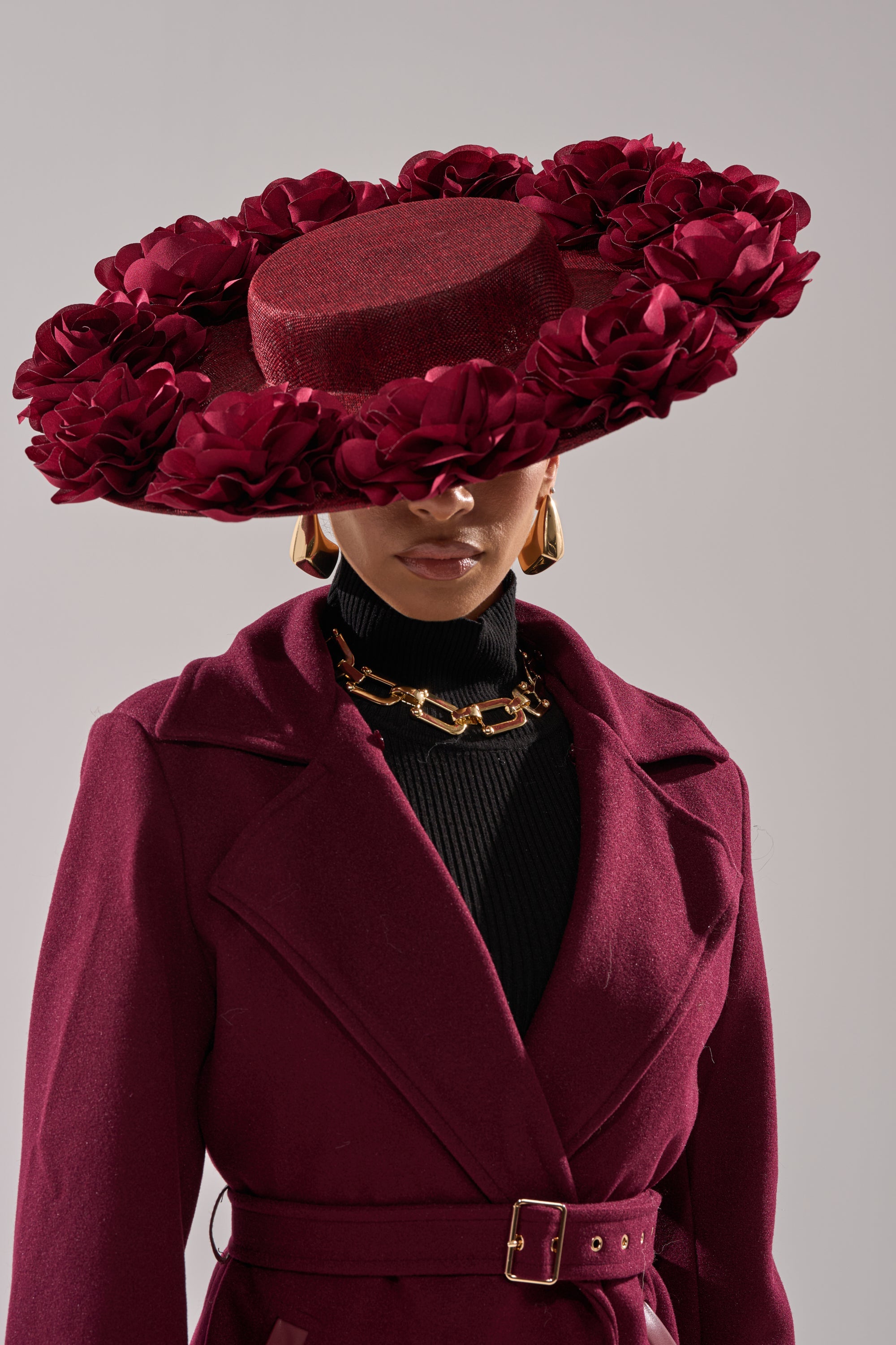 A person wears the ROSEY CLIP ON HAT—a burgundy wide-brimmed boater with 3D rose detailing—plus a matching belted coat, black turtleneck, chunky gold necklace, and gold earrings. The hat obscures their eyes. 0