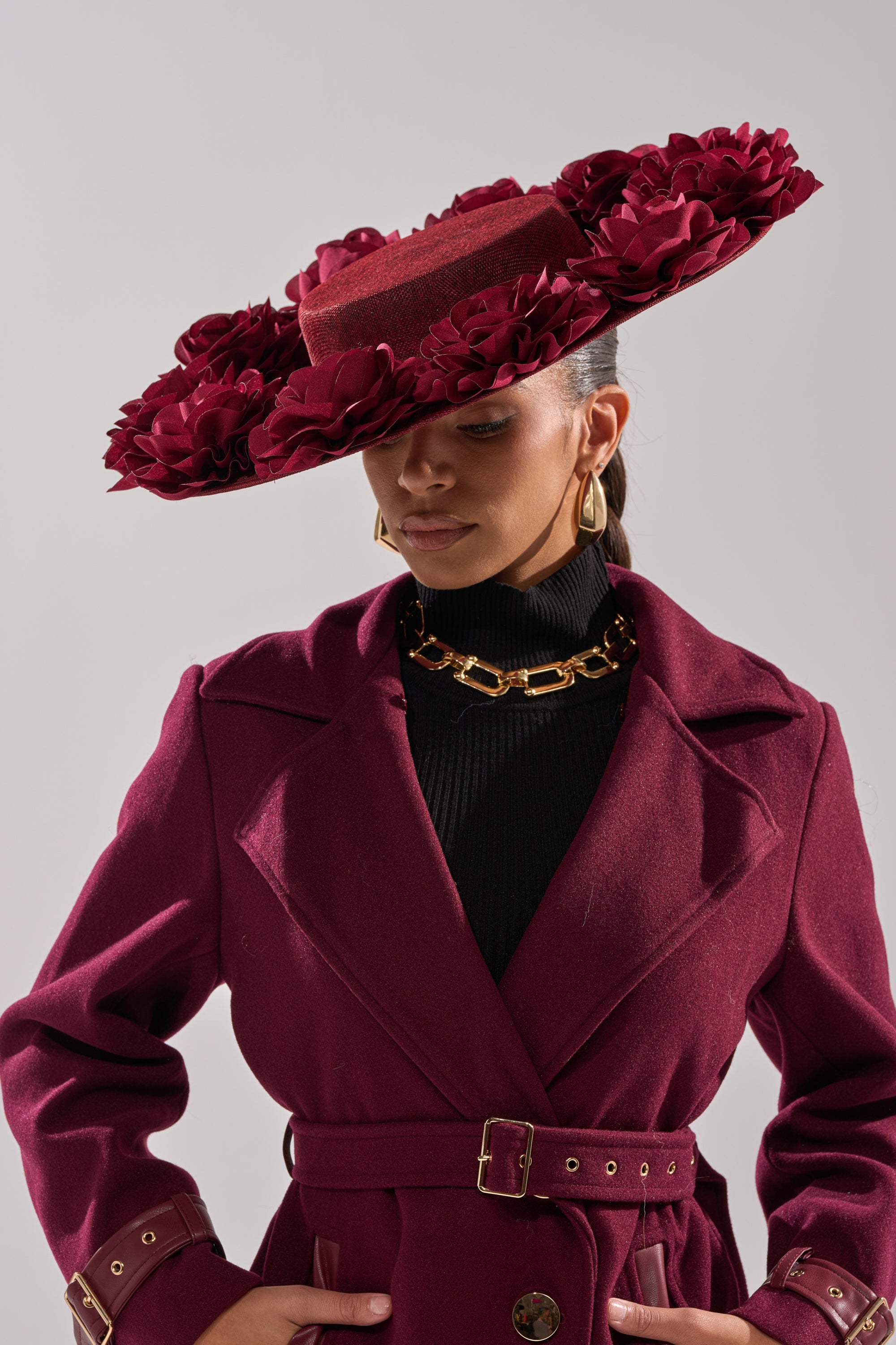 A woman wearing a deep burgundy ROSEY CLIP ON HAT with 3D rose detailing looks down, accessorizing her chic outfit with gold hoop earrings and a chunky gold chain necklace. 1