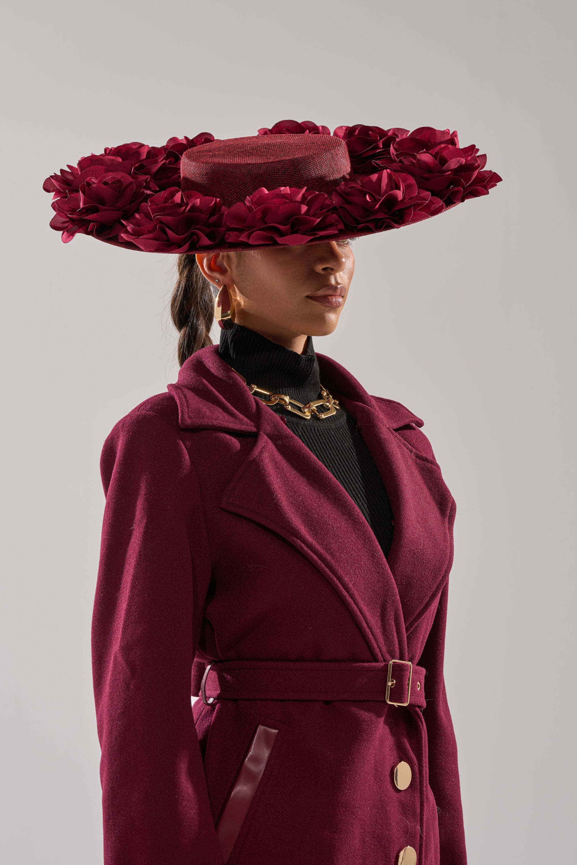 A woman in profile wears the ROSEY CLIP ON HAT, a wide-brimmed maroon boater with 3D rose details, paired with a matching maroon coat against a plain light background. 2