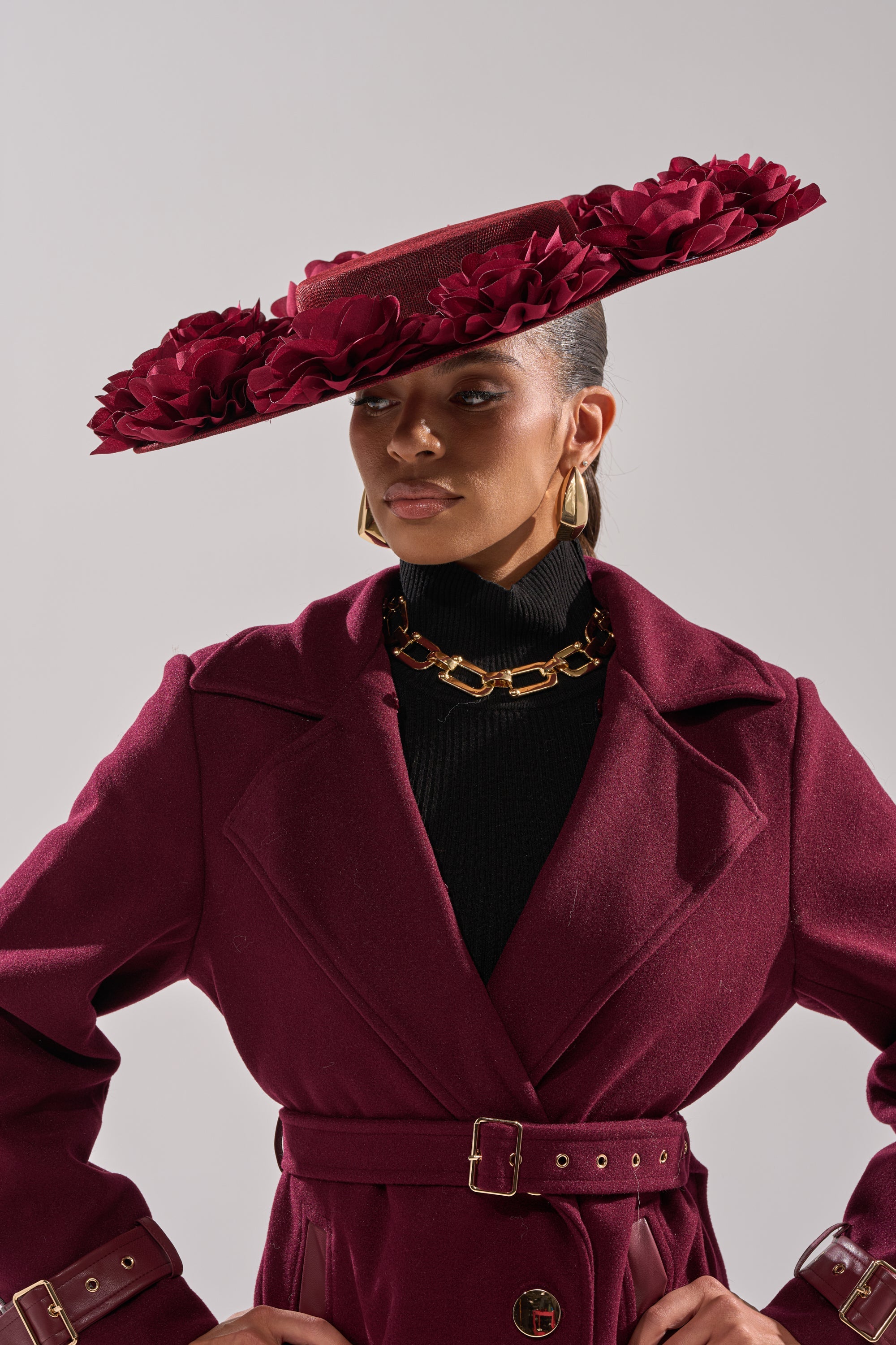 A woman wears a burgundy belted coat and the ROSEY CLIP ON HAT, a wide-brimmed piece adorned with 3D roses. Gold chain earrings and a necklace complete her look as she poses confidently against a neutral background. 3