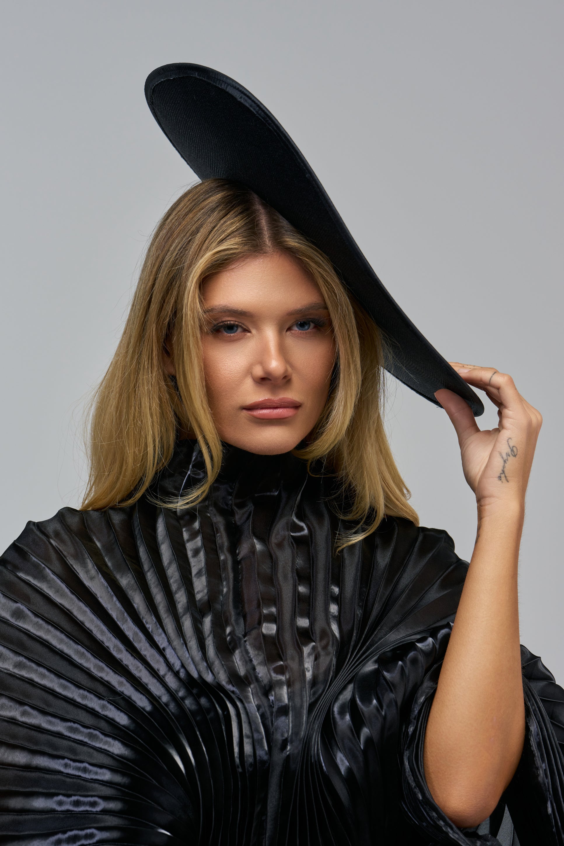 A person wearing an HBIC FASCINATOR, an oversized disc tilted over their forehead, and a black pleated high-neck top.