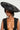 A person wearing the GIVING EDITORIAL HAT, an oversized wide-brimmed design in black, paired with a deep V-neck lace dress and large dangling cross earrings. The plain light-colored background accentuates the vintage glamor of this elegant and bold fashion statement.